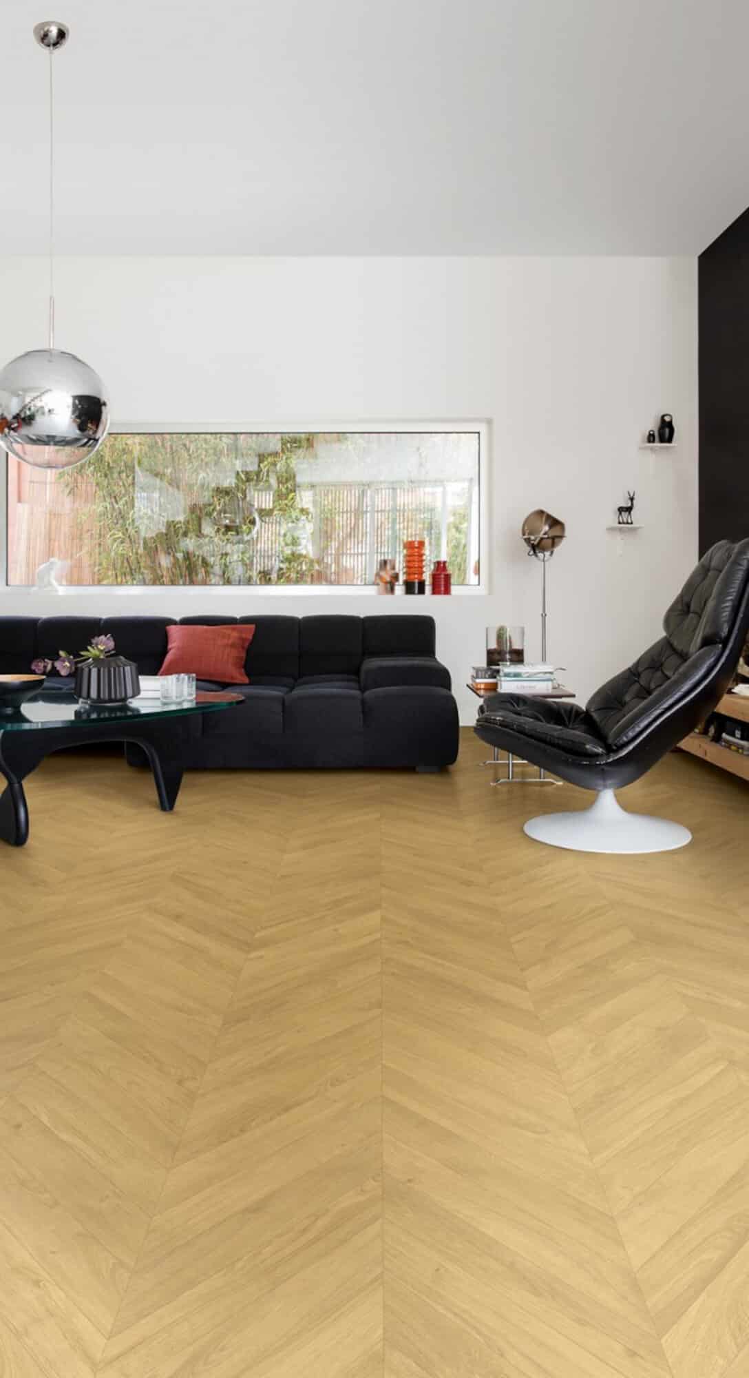 Ein modernes Wohnzimmer mit einem schwarzen Sofa, einem gläsernen Couchtisch und einem stilvollen schwarzen Sessel. Der Boden ist in einem hellen, hölzernen Fischgrätmuster gestaltet. An der Wand hängen dekorative Elemente, und ein großes Fenster lässt viel Tageslicht herein. Eine schimmernde, runde Deckenlampe ergänzt die Einrichtung.
