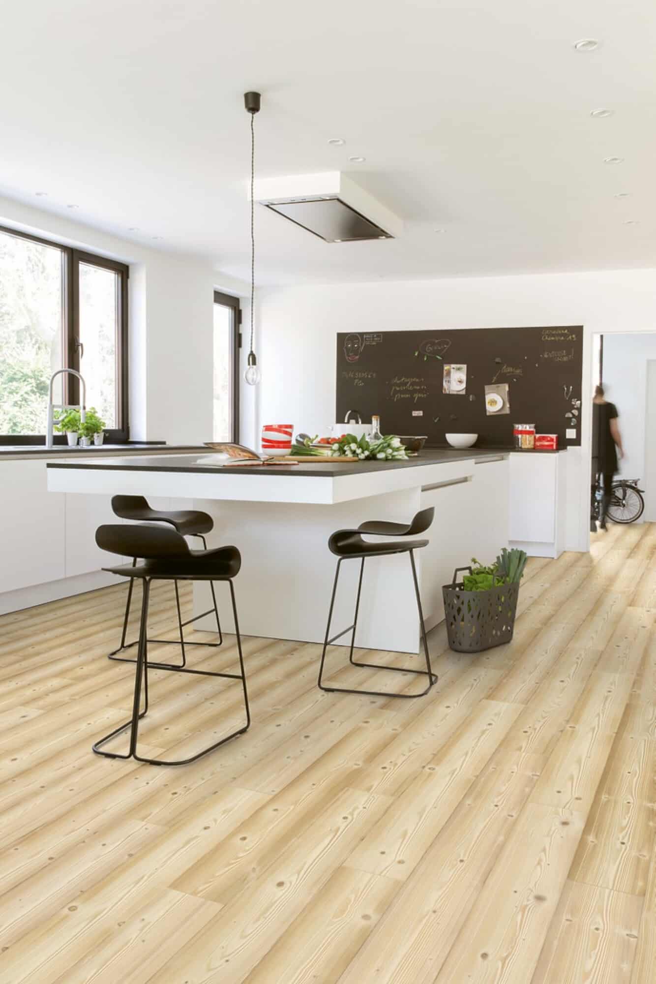 Moderne Küche mit minimalistischer Ausstattung, einem großen Kücheninseln und schwarzen Barhockern. Helle Wände und ein Holzboden sorgen für eine freundliche Atmosphäre. Im Hintergrund ist eine Tafel mit Notizen und Zeichnungen zu sehen.