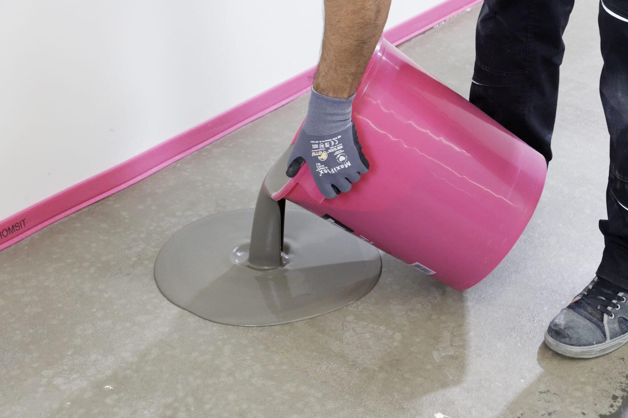 Eine Person gießt grauen Estrich aus einem pinken Eimer auf einen Betonboden, während sie Arbeitshandschuhe trägt. Im Hintergrund ist eine pinke Wandverkleidung zu sehen.