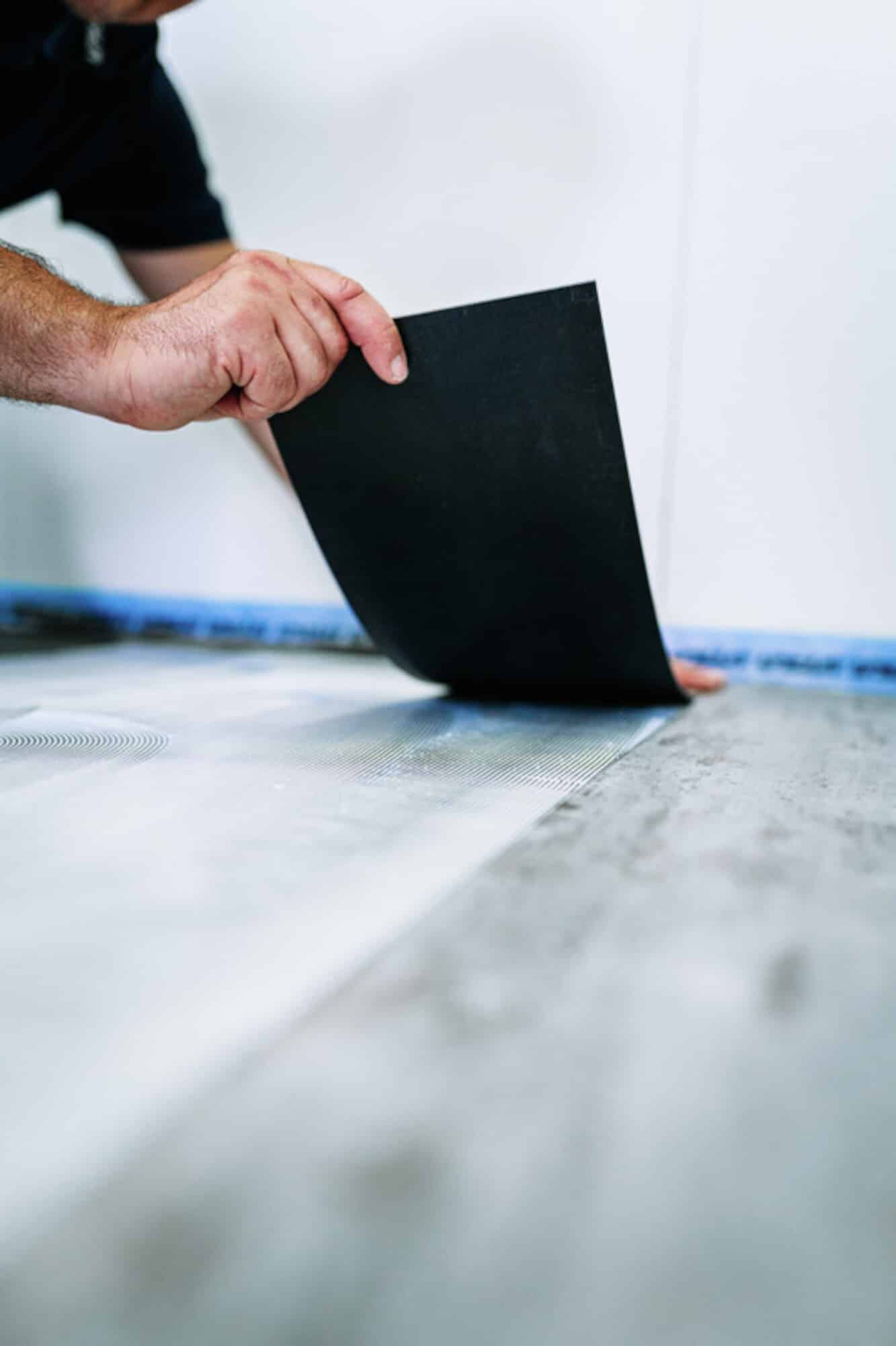 Eine Hand legt ein schwarzes Material auf einen grauen Boden, während im Hintergrund eine Wand zu sehen ist.