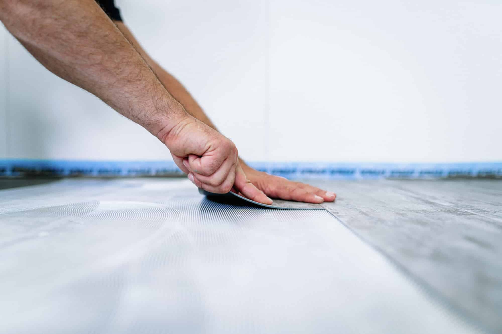 Eine Hand legt einen Bodenbelag auf einen Holzfußboden, während eine andere Hand das Material glattstreicht. Im Hintergrund ist eine Wand und ein blauer Streifen sichtbar.