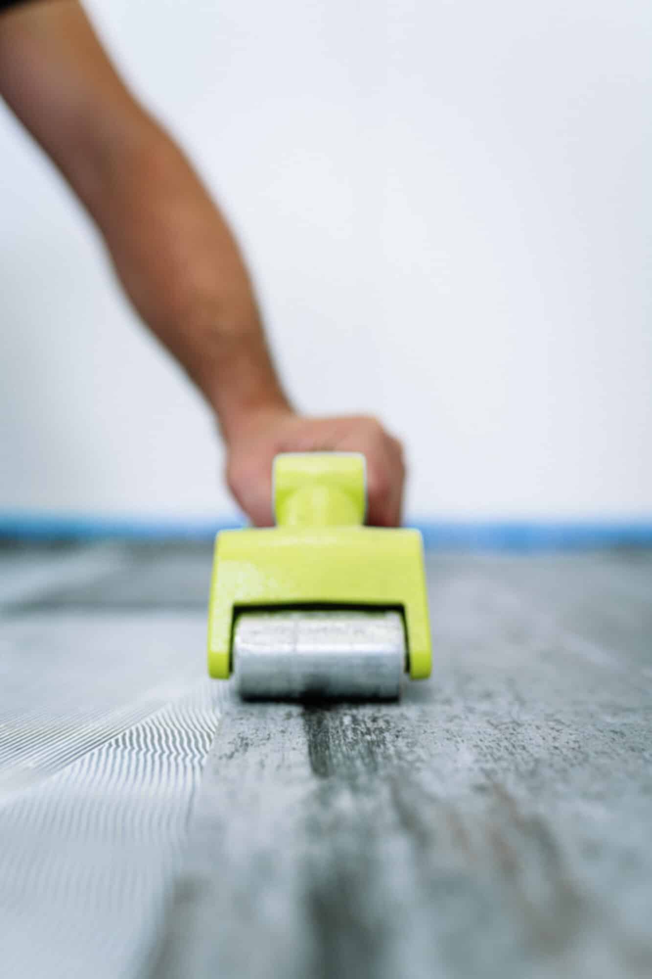 Ein Handwerker rollt mit einem grünen Werkzeug über einen Bodenbelag, während eine Hand im Vordergrund sichtbar ist. Der Hintergrund ist unscharf und zeigt eine weiße Wand sowie einen blauen Malerbandstreifen.