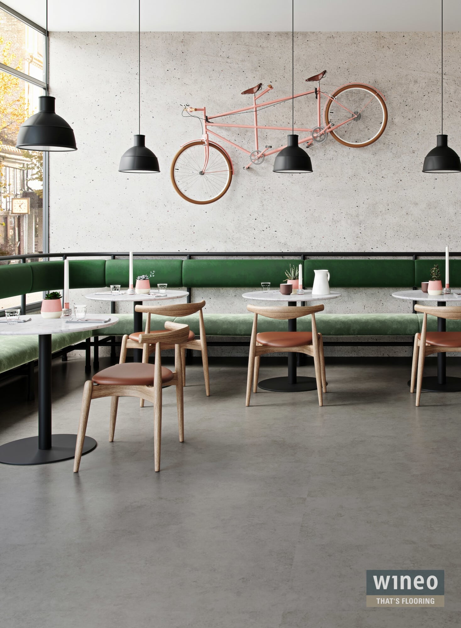 Ein modernes Café-Interieur mit grauen Wänden und einer grünen Bank. An den Wänden hängen schwarze Lampen, während ein pinkfarbenes Fahrrad als Dekoration dient. Es gibt runde Tische mit weißen Tischplatten und Holzstühlen mit braunen Sitzflächen. Kleine Pflanzen und Kerzen sind auf den Tischen platziert.