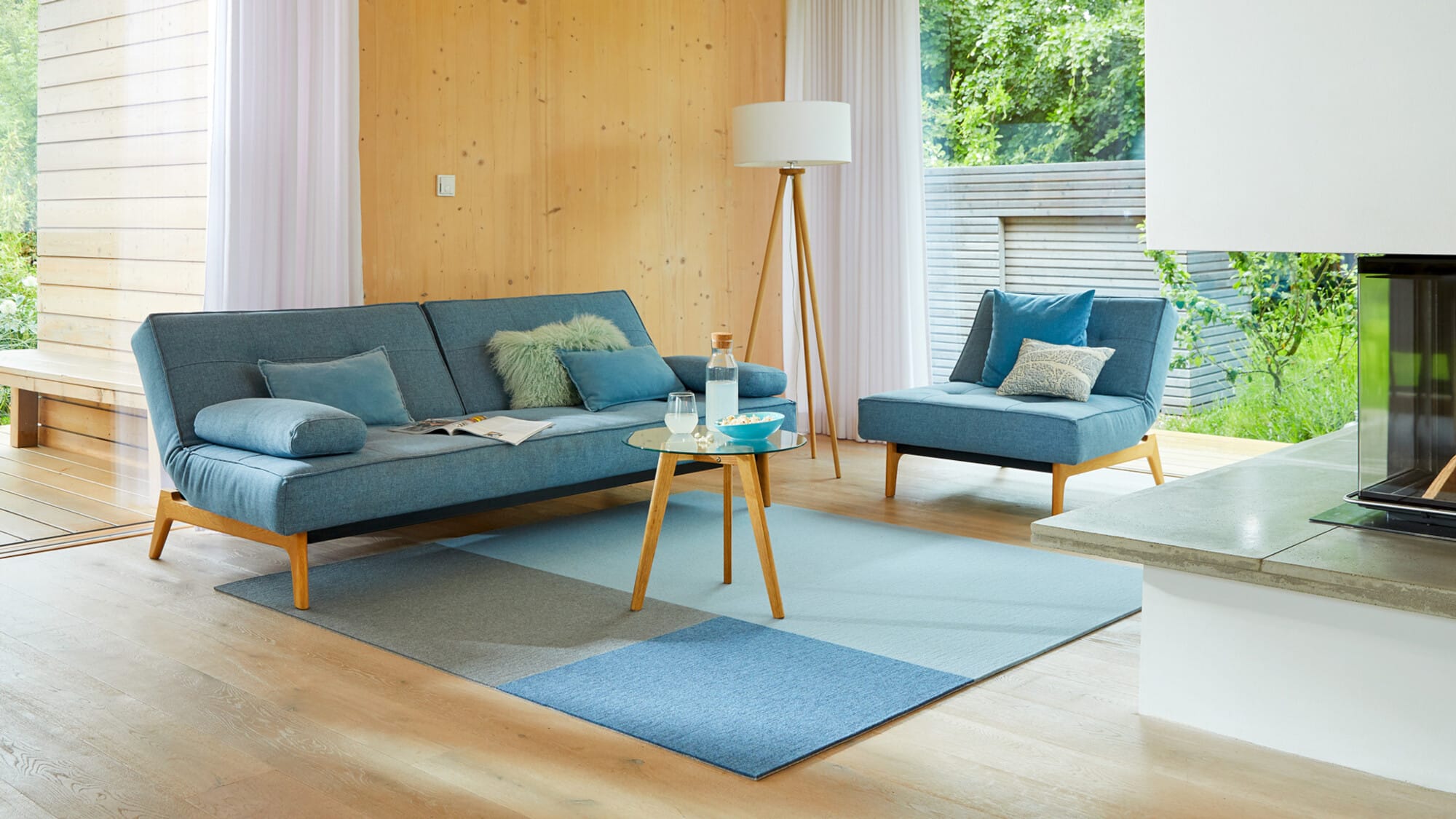 Ein modernes Wohnzimmer mit einem blauen Sofa und einem passenden Sessel, umgeben von Holzverkleidung und großen Fenstern, die viel Tageslicht hereinlassen. Ein runder Glastisch steht in der Mitte auf einem farblich abgestimmten Teppich. Dekorative Kissen und eine Stehlampe ergänzen die gemütliche Atmosphäre.