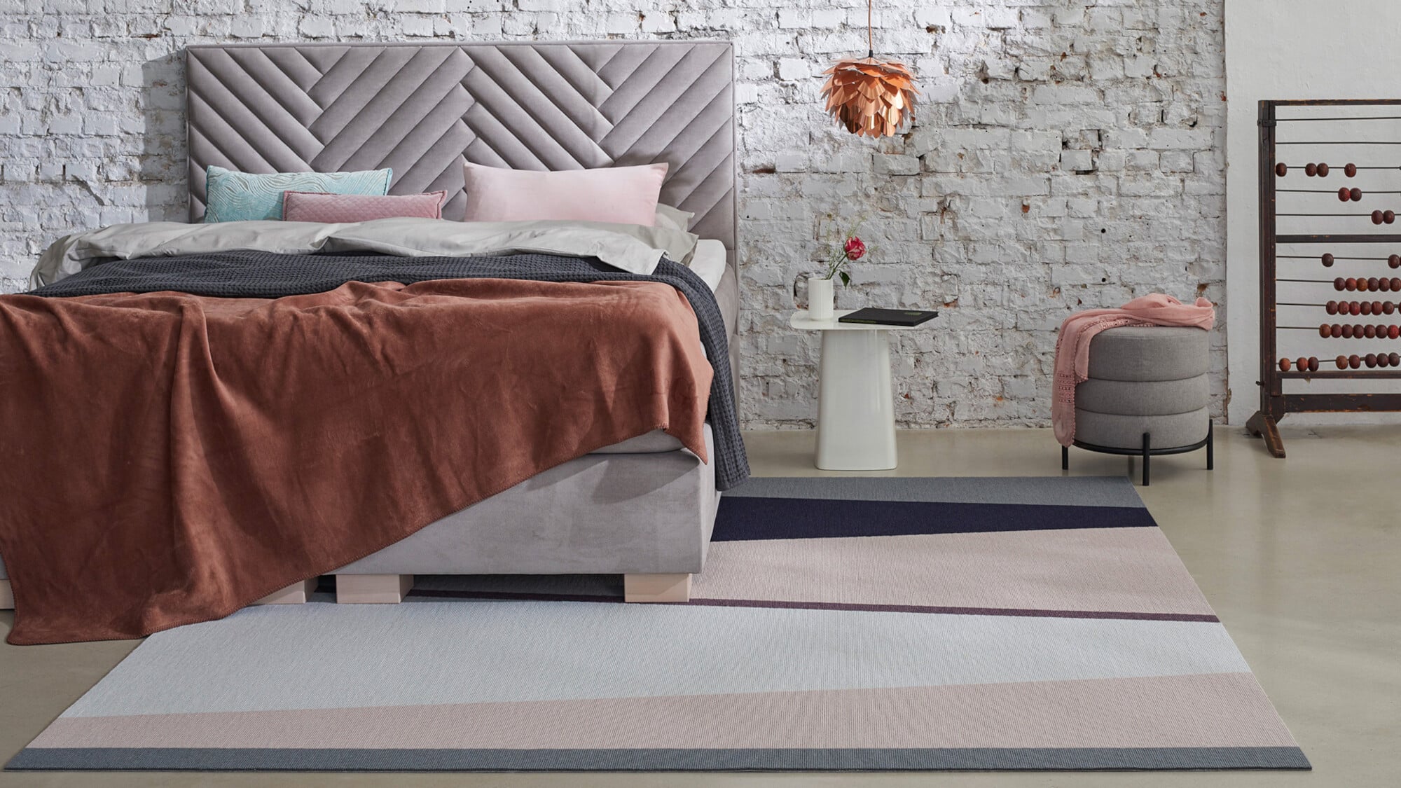 Ein modernes Schlafzimmer mit einem gepolsterten Bett und einer gesteppten Kopfstütze. Die Bettwäsche ist in sanften Farben gehalten, mit einer braunen Decke und verschiedenen Kissen. Neben dem Bett steht ein minimalistischer Nachttisch mit einer Blume und einem Notizblock. Auf dem Boden liegt ein gestreifter Teppich in harmonischen Farbtönen. An der Wand hängt eine kreative Lampe, und im Hintergrund ist eine alte Wand mit Ziegelsteinen zu