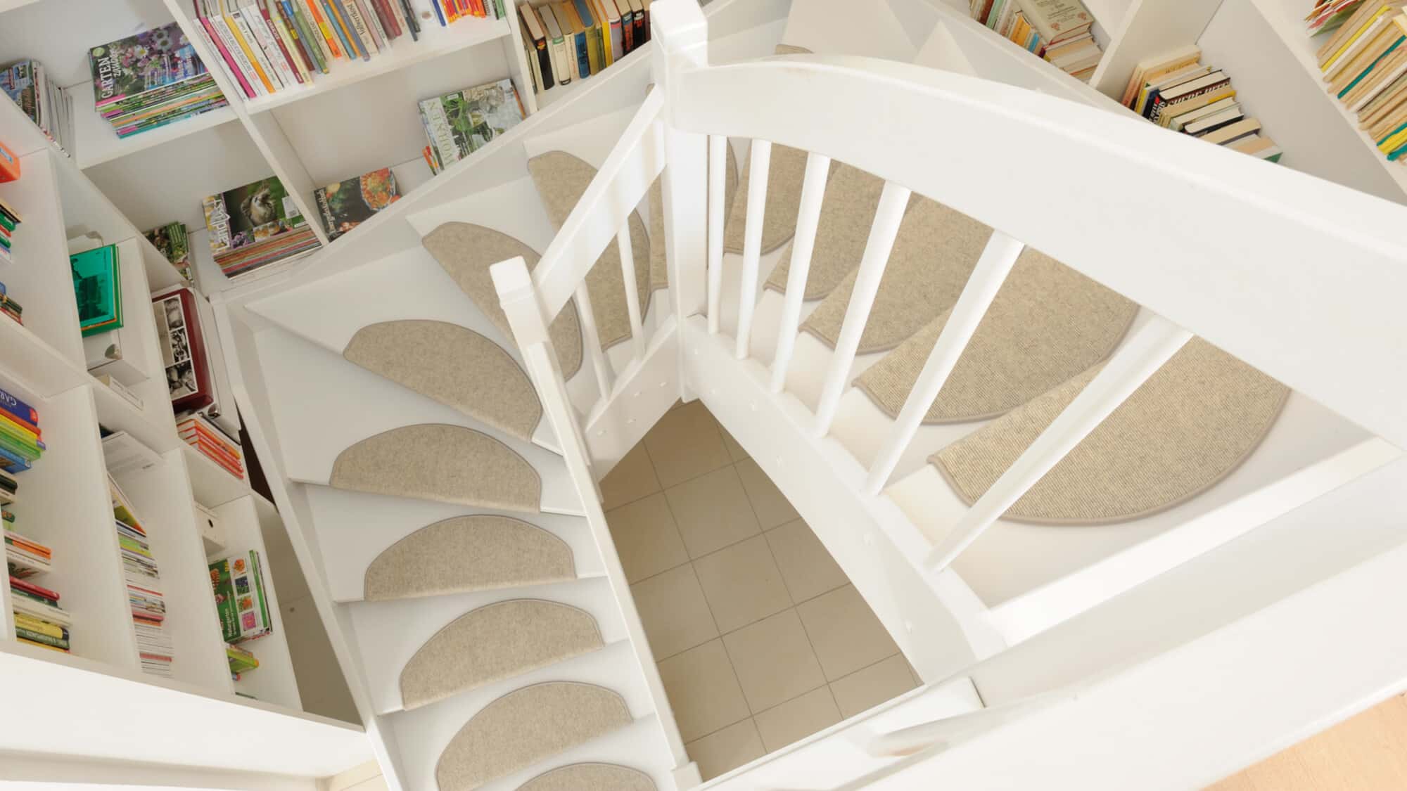 Eine spiralförmige Treppe mit weißen Stufen und runden, beigen Teppichauflagen, umgeben von Bücherregalen, die mit verschiedenen Büchern gefüllt sind. Der Blick ist von oben auf die Treppe gerichtet, die in einen hellen Raum führt.