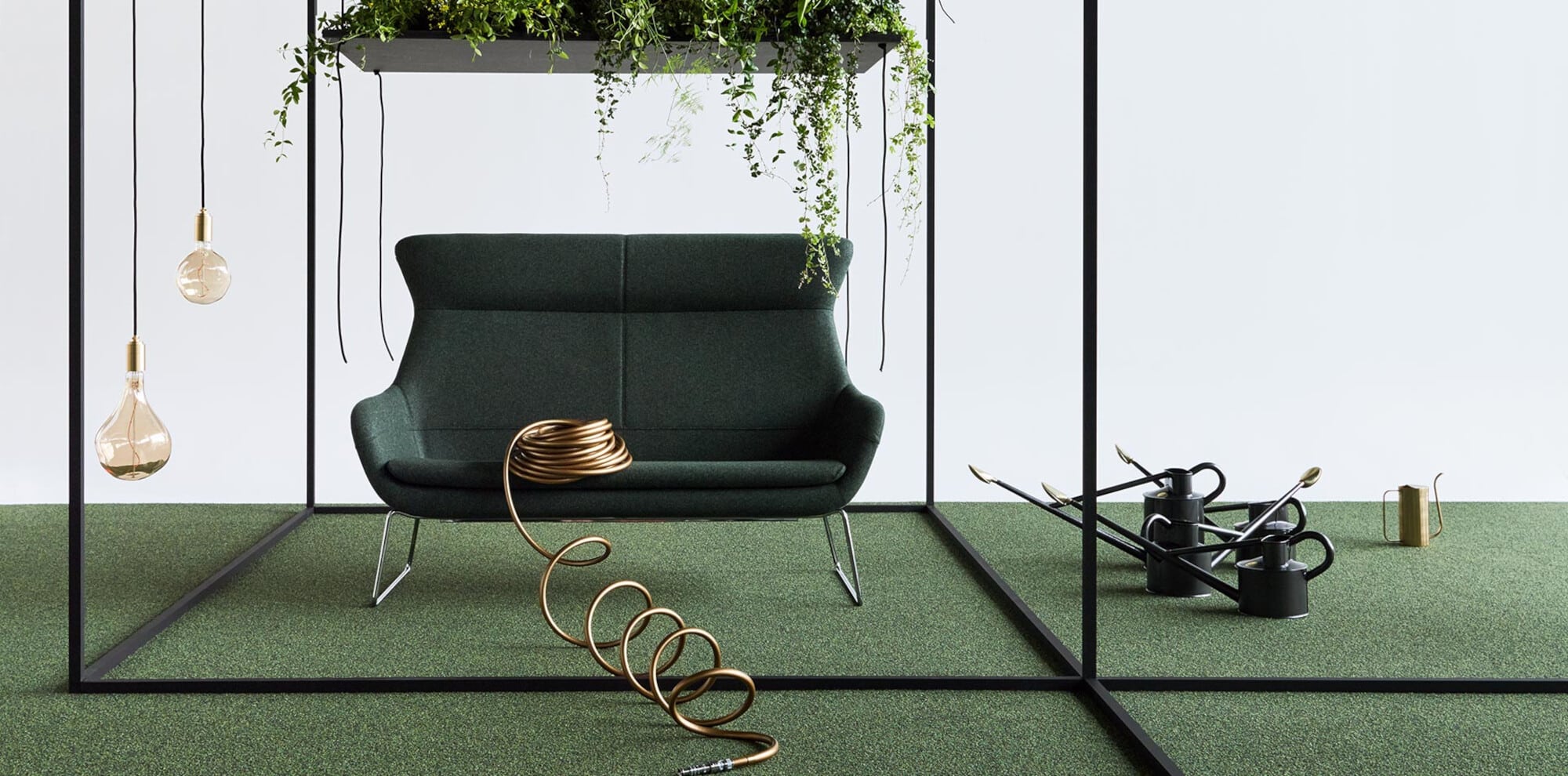 Ein stilvolles Wohnzimmer mit einem grünen Sofa, umgeben von Pflanzen und hängenden Lampen. Im Vordergrund liegt ein goldfarbener Schlauch, während im Hintergrund Gießkanne und Werkzeuge sichtbar sind. Der Boden ist mit grünem Teppich bedeckt, was eine frische, natürliche Atmosphäre schafft.