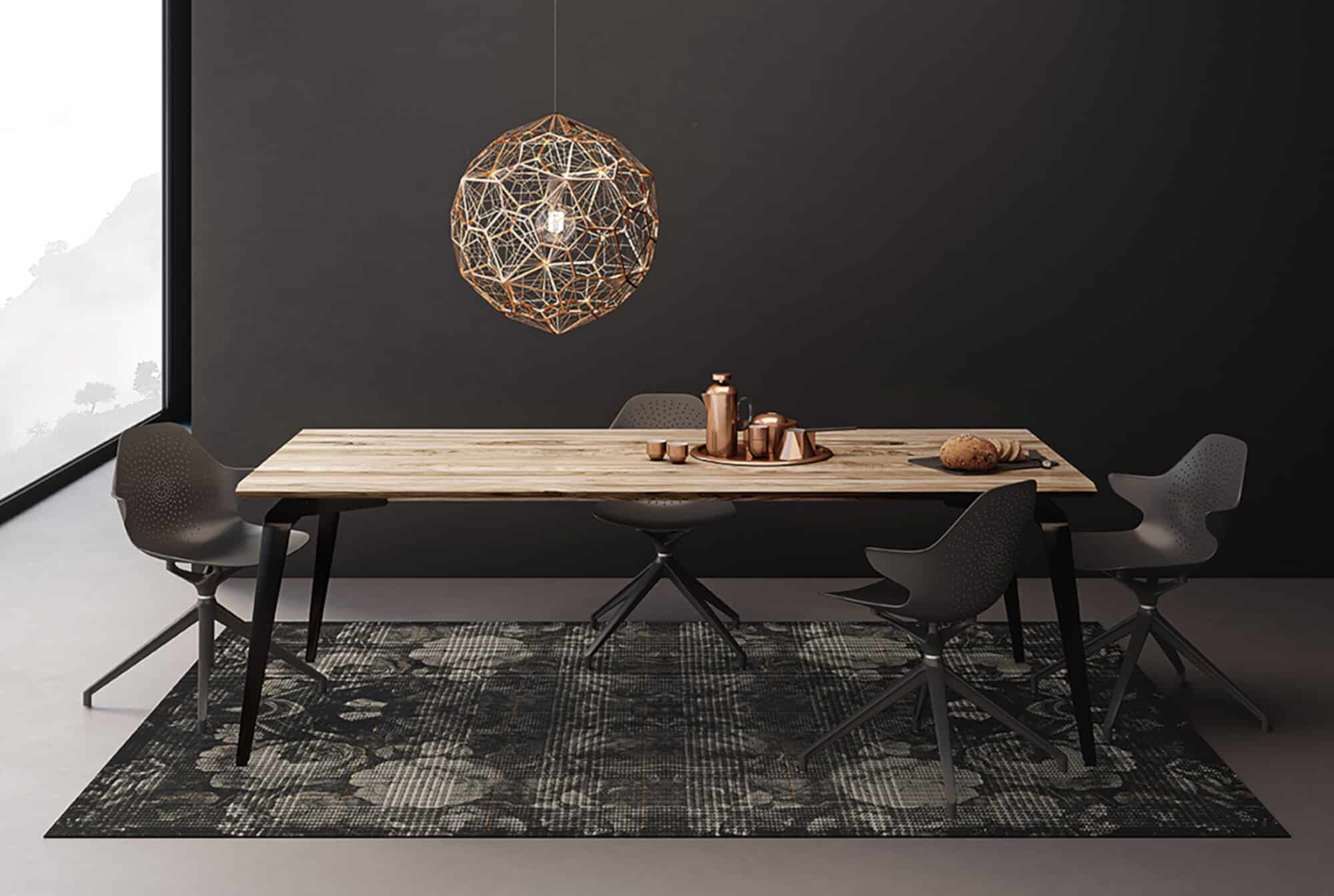 Ein modern eingerichteter Essbereich mit einem langen Holztisch und stilvollen schwarzen Stühlen. Über dem Tisch hängt eine auffällige, geometrische Pendelleuchte. Auf dem Tisch stehen eine Kaffeekanne, Tassen und ein Brot, während ein gemusterter Teppich den Raum ergänzt. Die Wände sind dunkel, was einen eleganten Kontrast zur hellen Tischoberfläche bietet.