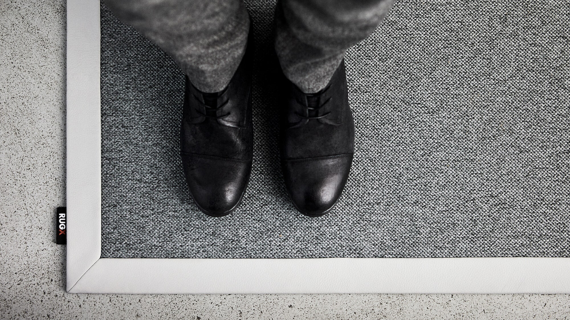 Schwarze Schuhe stehen auf einem grauen Teppich mit einem weißen Rand. Im Hintergrund ist ein glatter, heller Boden zu sehen.