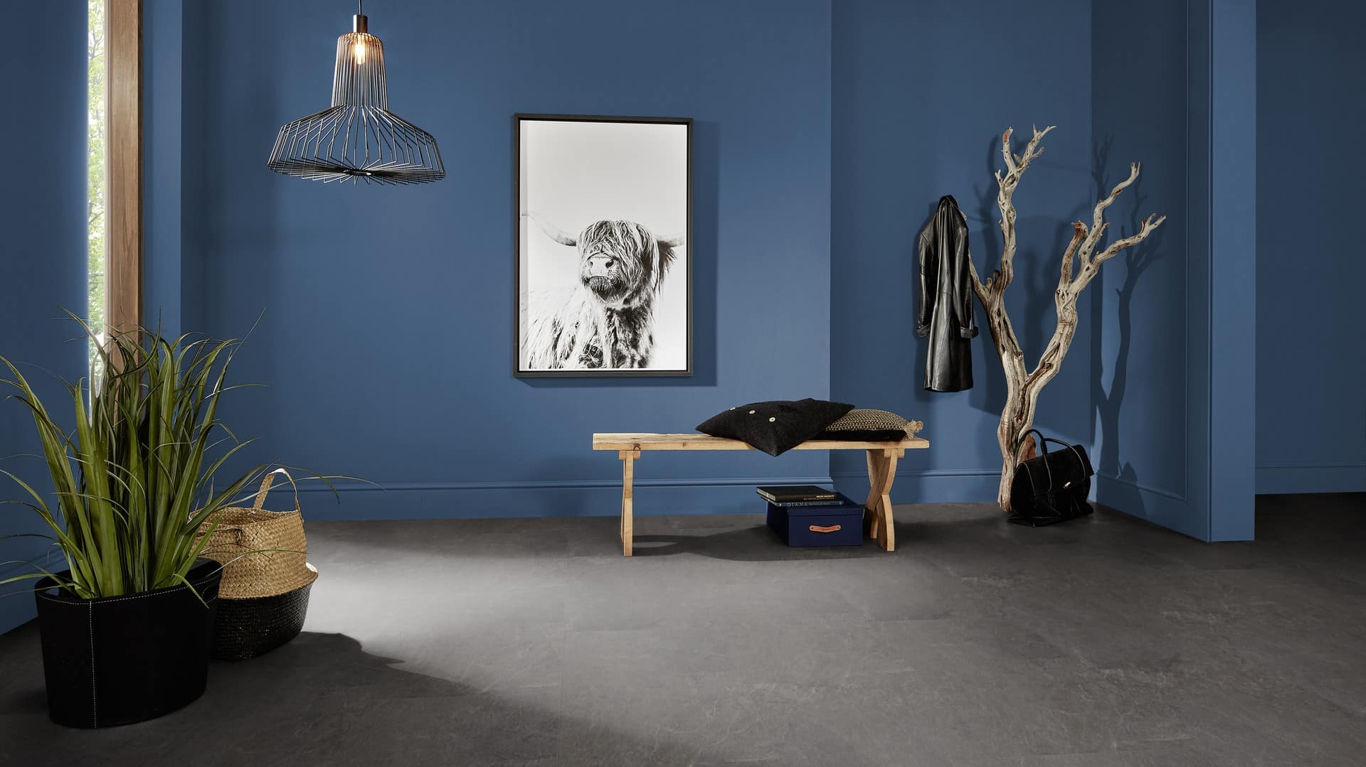Ein moderner Eingangsbereich mit blauen Wänden, einer stylischen Hängeleuchte, einer Holzbank mit Kissen, einem schwarzen und einem geflochtenen Korb, sowie einem dekorativen Baumstamm und einem schwarzen Mantel, der an der Wand hängt. Ein Schwarz-Weiß-Bild eines Hochlandrinds ist an der Wand angebracht.