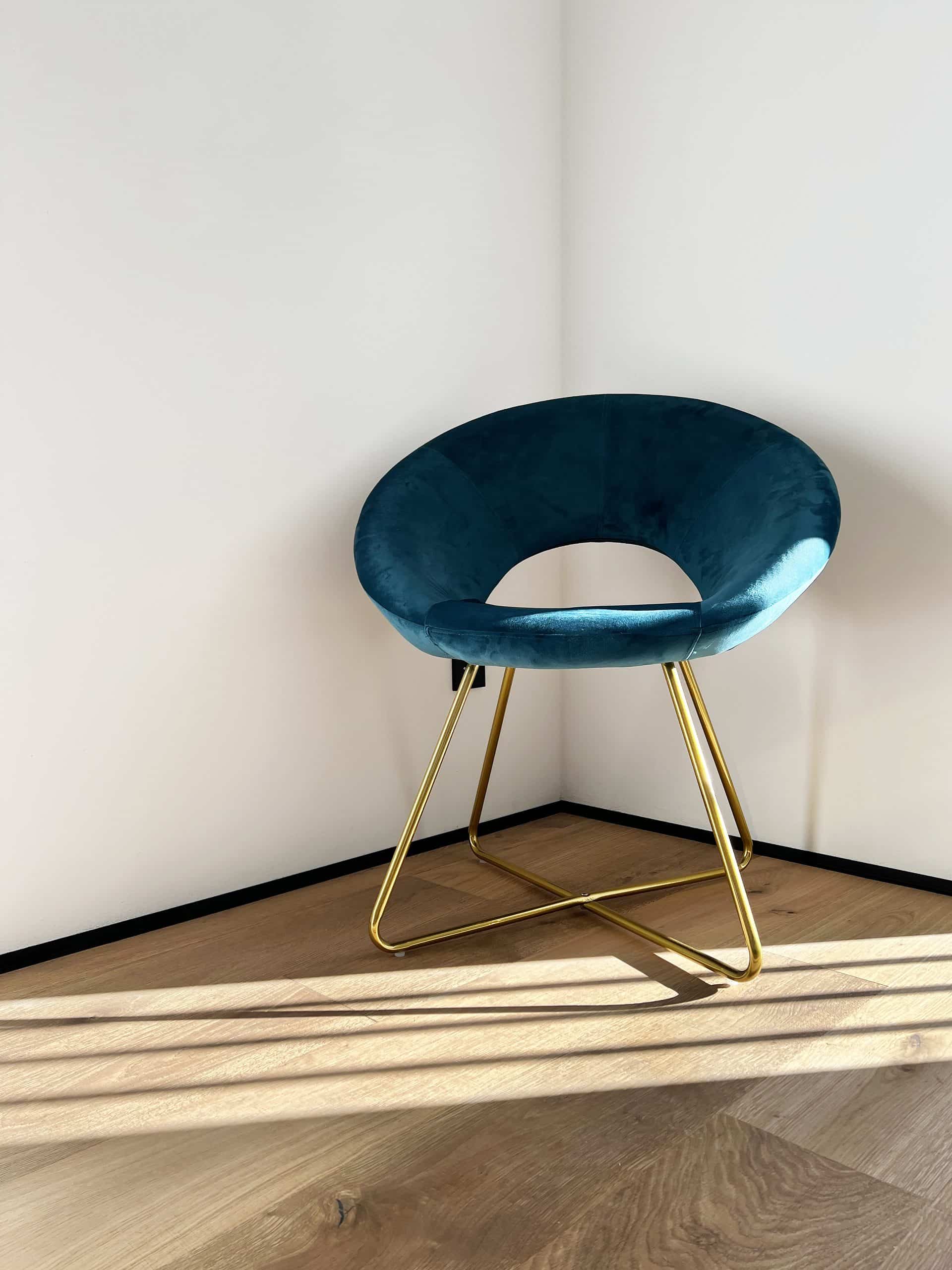 Ein eleganter, runder Sessel mit einem samtigen, blauen Bezug steht in einer Ecke eines Raumes. Der Sessel hat ein modernes Gestell aus goldfarbenem Metall, und das Licht fällt sanft auf den Boden aus Holz, wodurch interessante Schattenmuster entstehen.