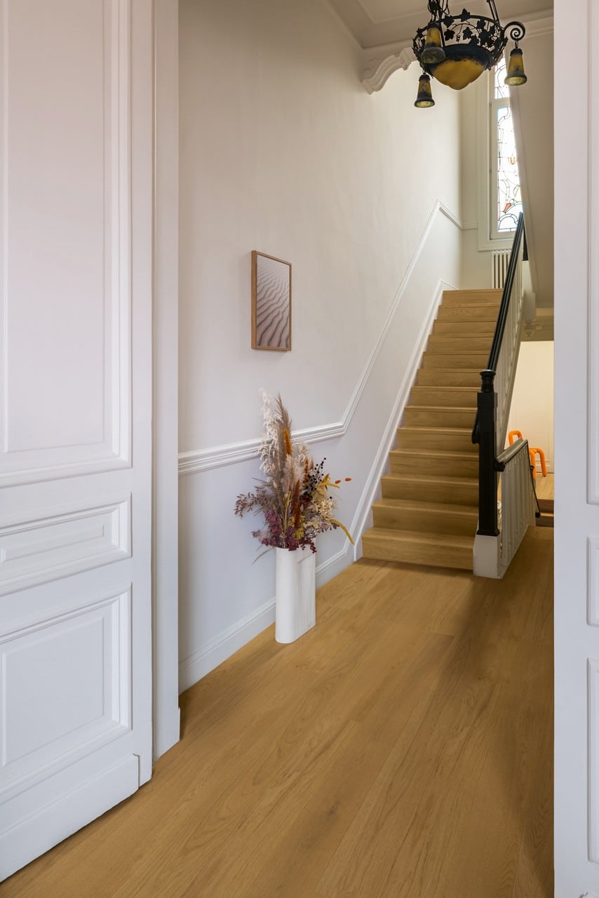 Ein heller Flur mit einer Holztreppe, die nach oben führt. An der Wand hängt ein gerahmtes Bild, und ein eleganter Blumentopf mit Trockenblumen steht auf dem Boden. Die Wände sind in sanften Farben gehalten, und eine dekorative Deckenlampe beleuchtet den Raum.