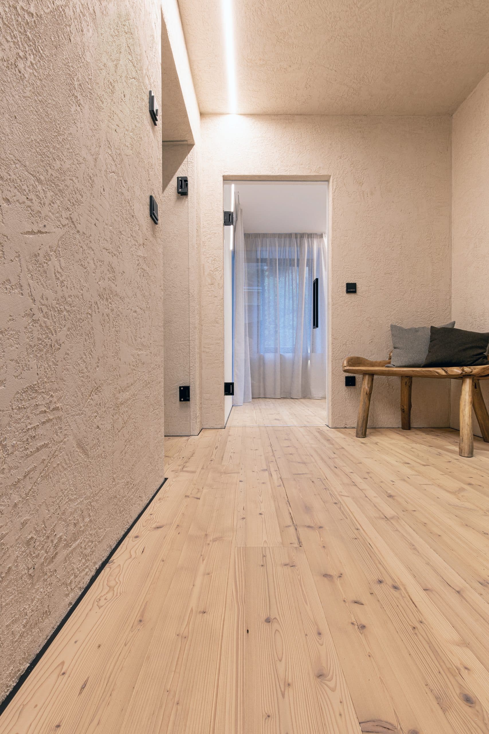 Ein heller Flur mit strukturierten Wänden in Beige, einem hellen Holzboden und einem rustikalen Holzbank. Im Hintergrund sind Vorhänge und ein Fenster sichtbar, das Tageslicht hereinlässt.