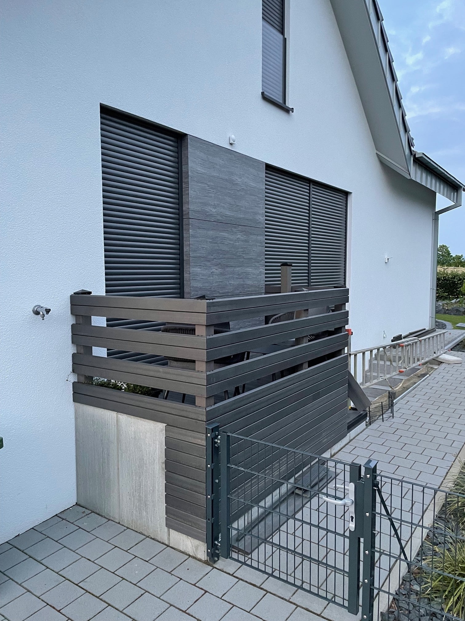 Ein moderner Eingang zu einem Wohnhaus mit einem kleinen, überdachten Bereich, der von einer Holzverkleidung umgeben ist. Der Boden besteht aus grauen Pflastersteinen, und es gibt ein Gittertor, das den Zugang zum Bereich ermöglicht. Die Wand des Hauses ist weiß, und es sind Rollläden an den Fenstern sichtbar.