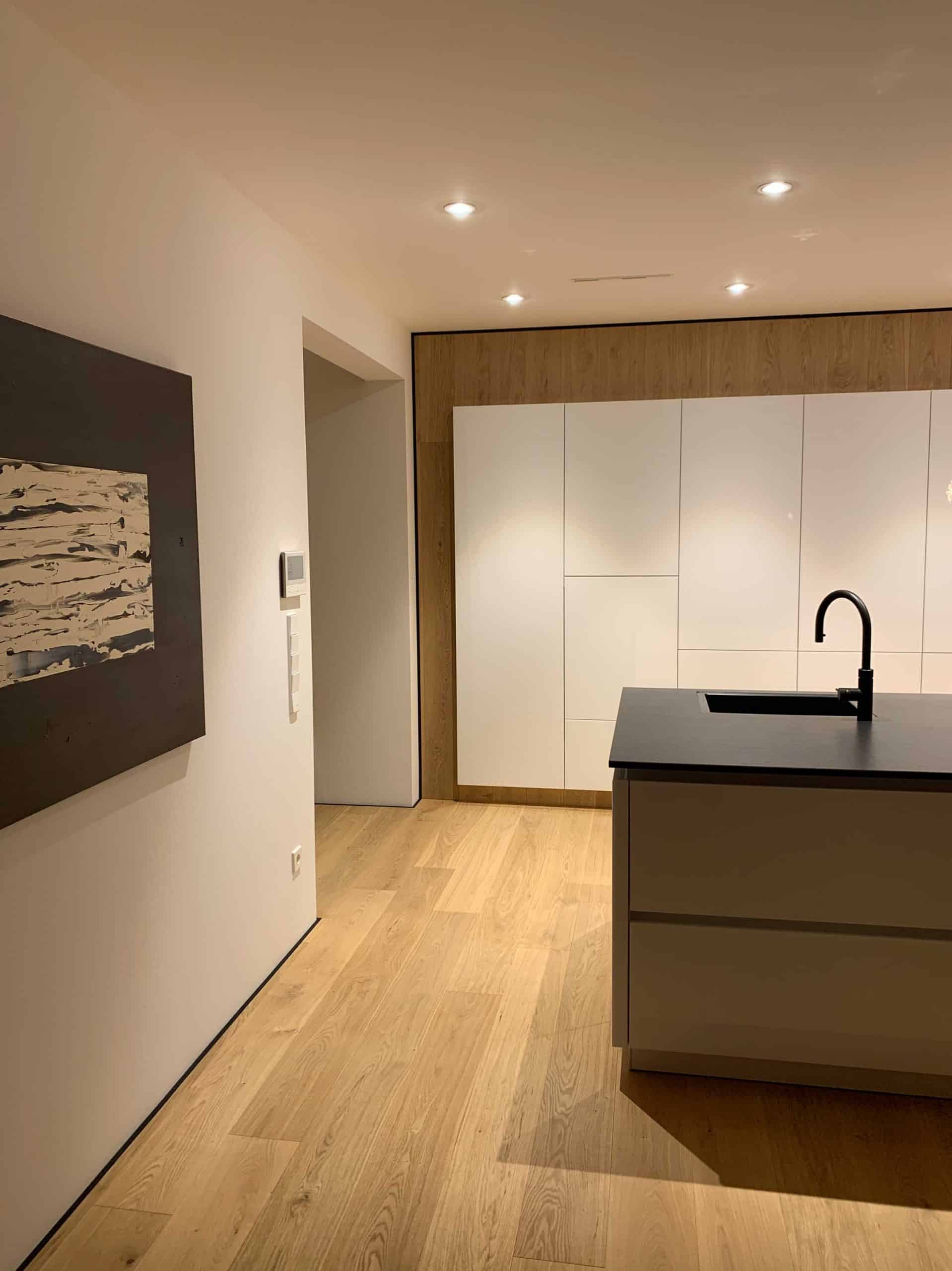 Moderne Küche mit Holzfußboden, weißen Küchenschränken und einer dunklen Kücheninsel. An der Wand hängt ein abstraktes Kunstwerk. Deckenbeleuchtung sorgt für eine helle Atmosphäre.