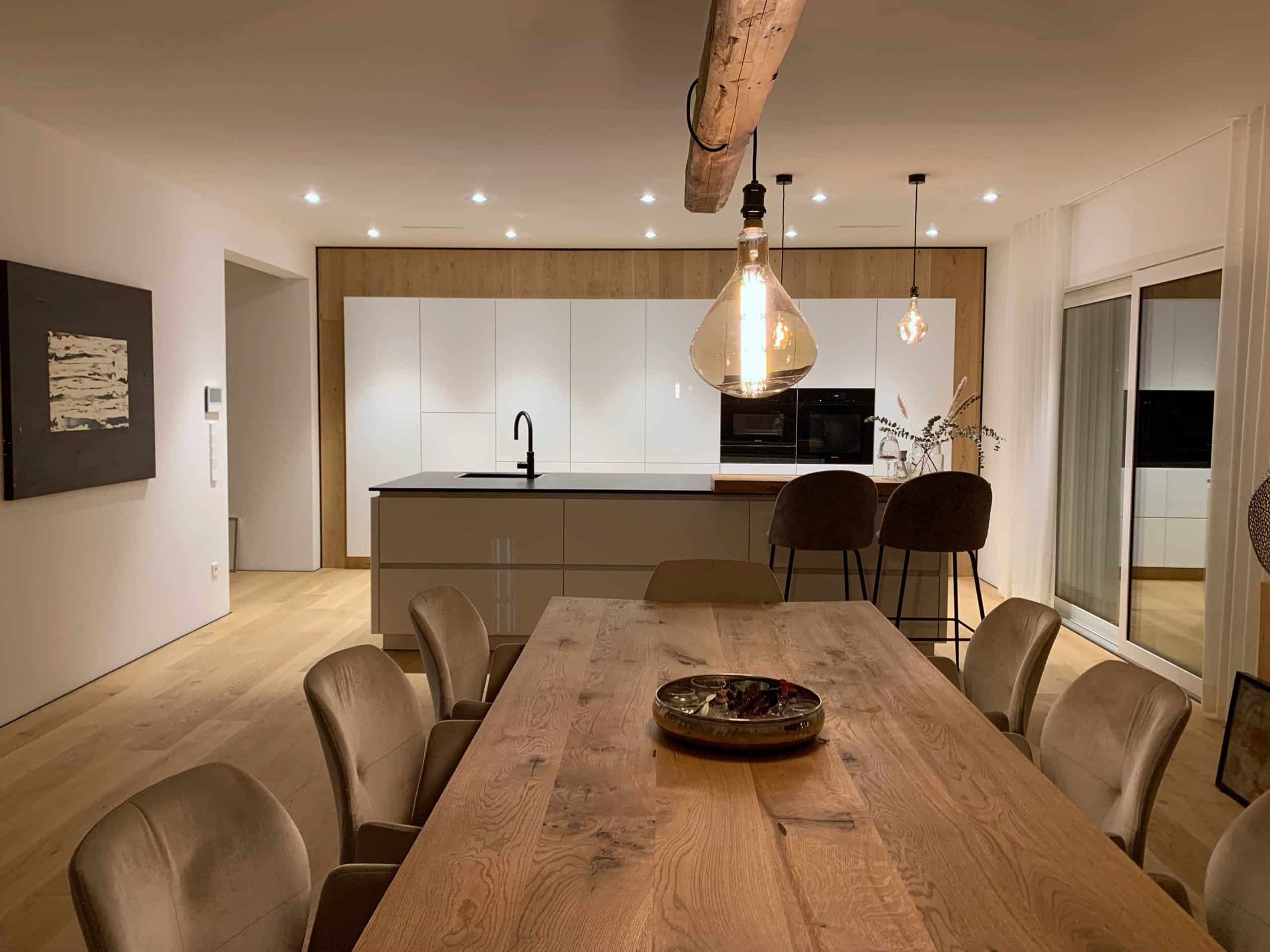 Moderne, helle Küche mit weißen Schränken, einer dunklen Kücheninsel und eleganten Barhockern. Im Vordergrund ein großer Holztisch mit dekorativem Geschirr. Große Fenster sorgen für viel Tageslicht.