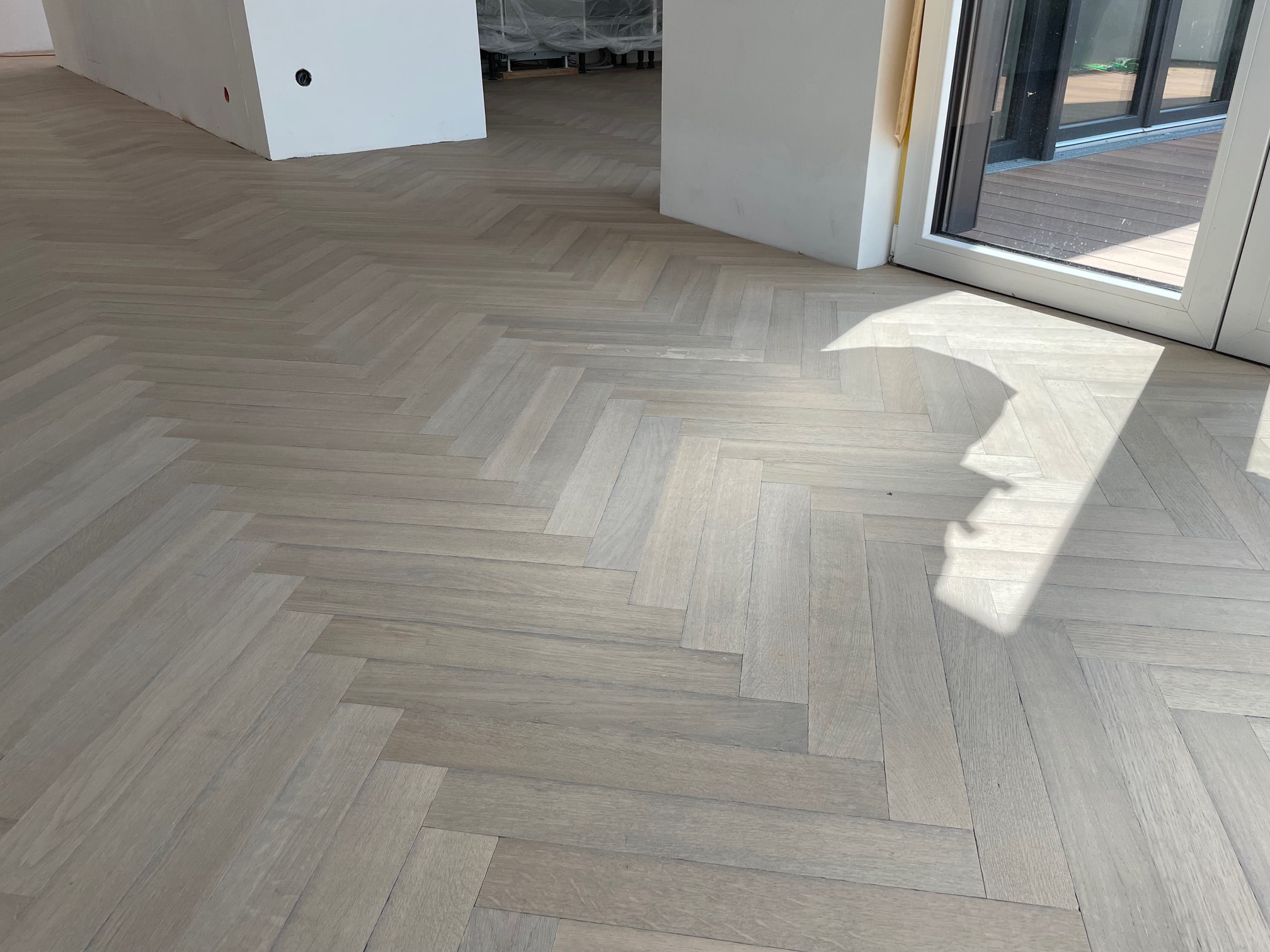 Der Boden eines Raumes mit hellen, schräg verlegten Holzplanken, die ein Fischgrätenmuster bilden. Das Licht fällt durch eine große Fensterfront und erzeugt Schatten auf dem Boden. Im Hintergrund sind noch nicht vollständig eingerichtete Wände und Möbel zu sehen.
