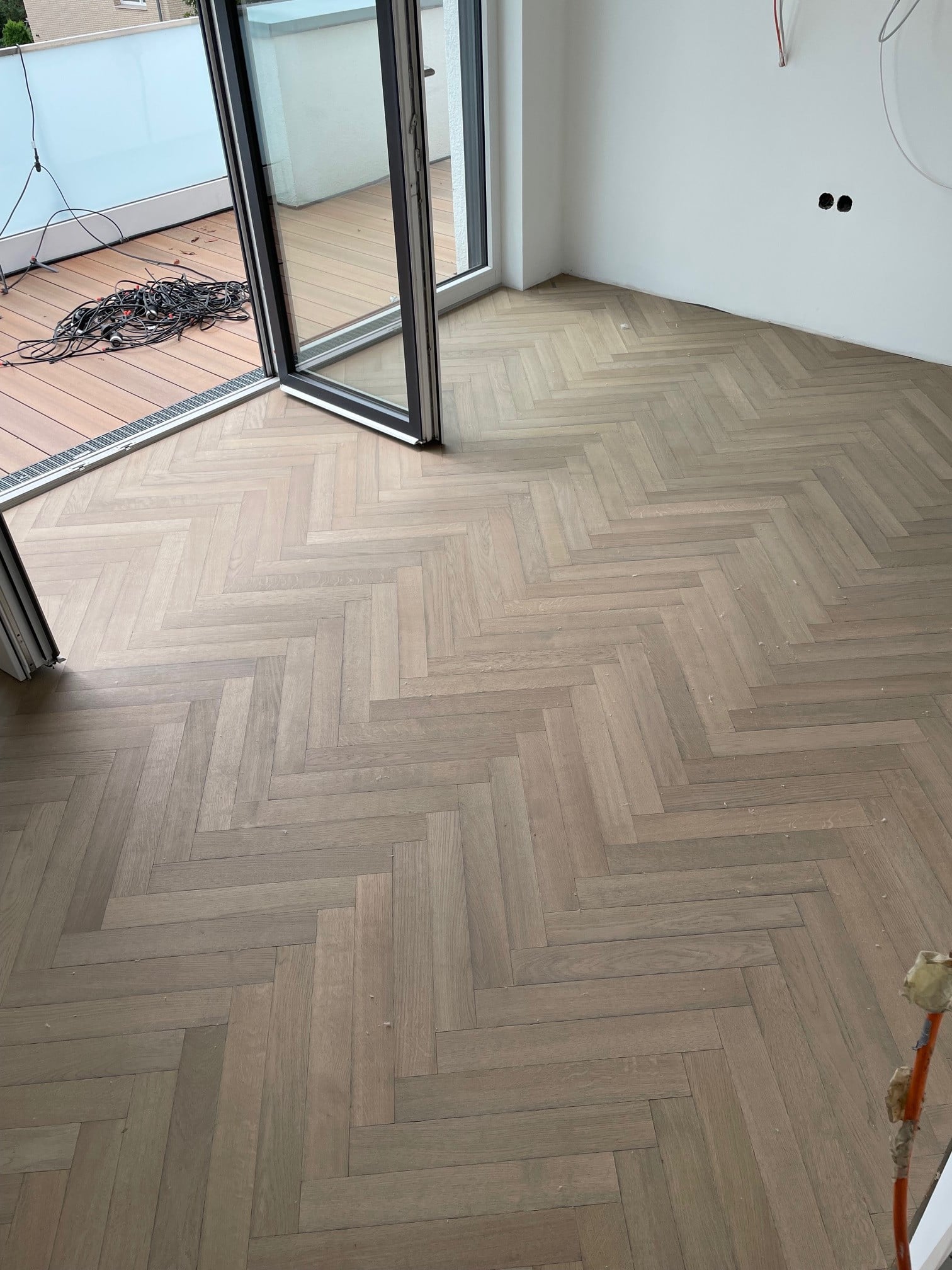 Der Boden eines Raumes mit Parkett in Fischgrätmuster, hellen Holztönen und einem großen Fenster, das auf einen Balkon führt. Kabel liegen auf dem Boden, und die Wände sind noch unvollständig.