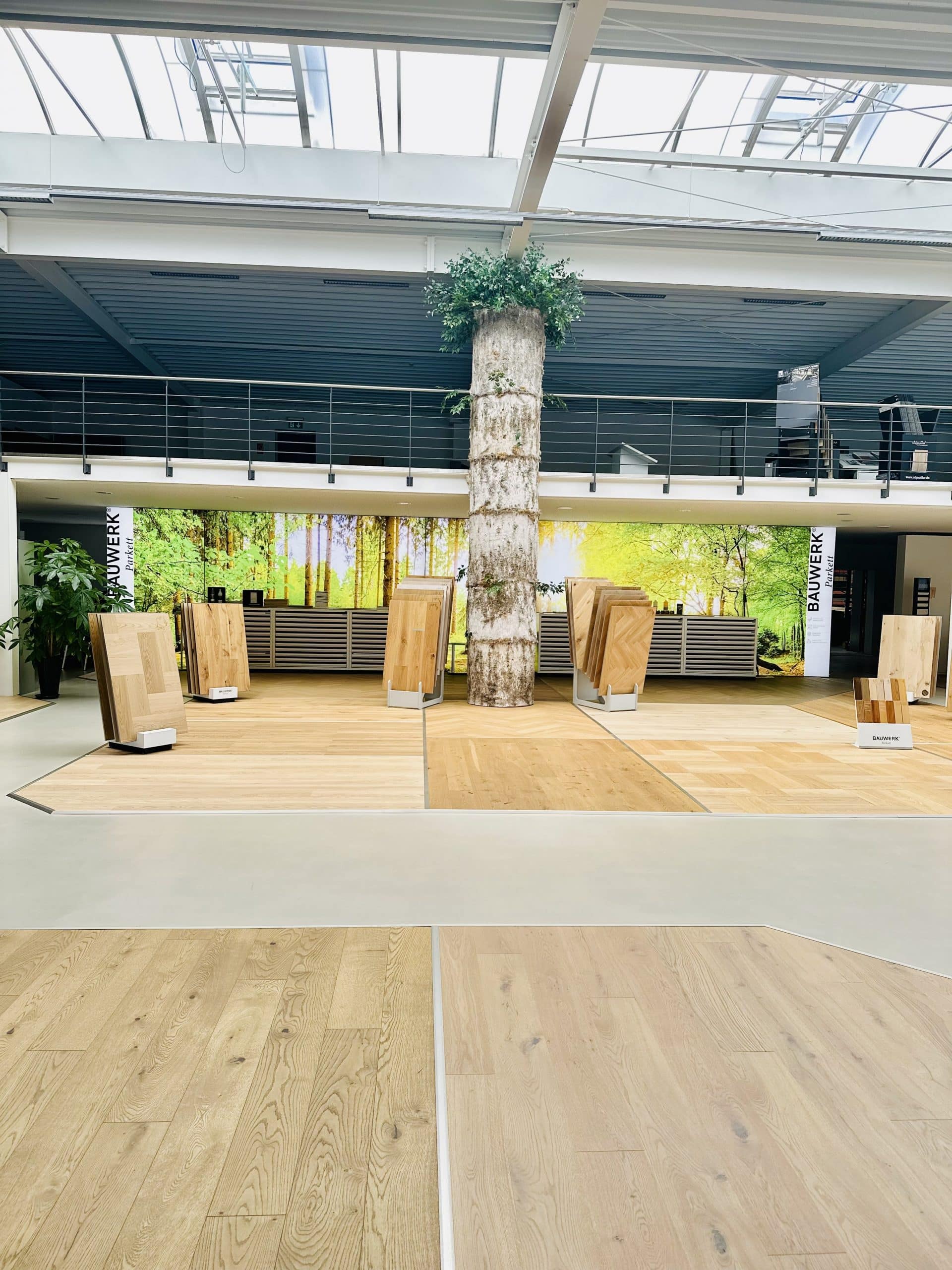 Ein moderner Ausstellungsraum mit Holzfußböden und Holzmustern, die auf Podesten präsentiert werden. In der Mitte steht ein Baumstamm, umgeben von Pflanzen. Im Hintergrund ist eine große Wand mit einem Waldmotiv zu sehen, und das Licht fällt durch ein Glasdach.