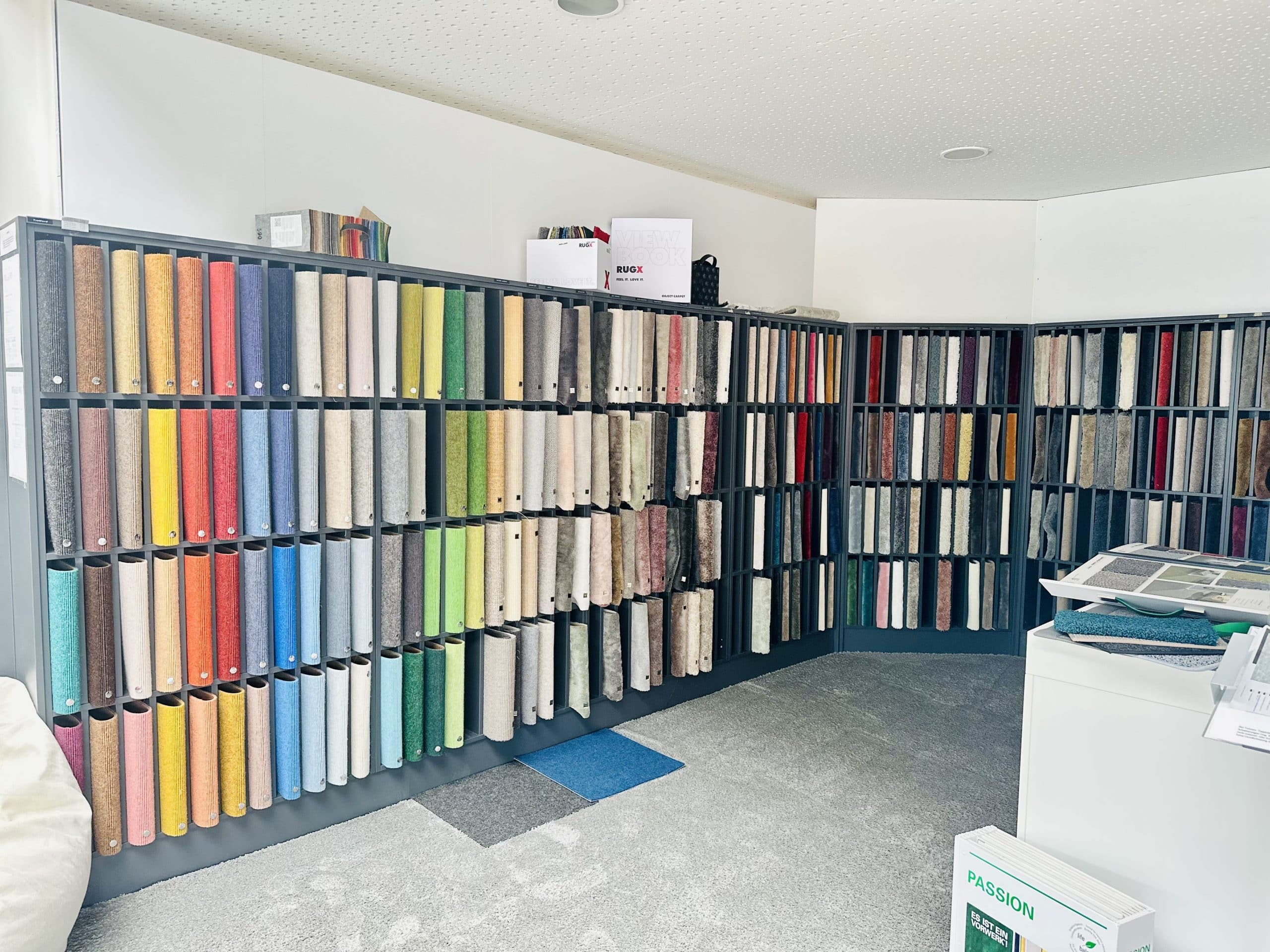 Ein Raum mit Regalen, die eine Vielzahl von Teppichmustern in unterschiedlichen Farben und Texturen zeigen. Die Wände sind mit Farbmusterrollen gefüllt, während der Boden mit grauem Teppich ausgelegt ist. Auf einem Tisch liegen einige Informationsblätter und Muster.