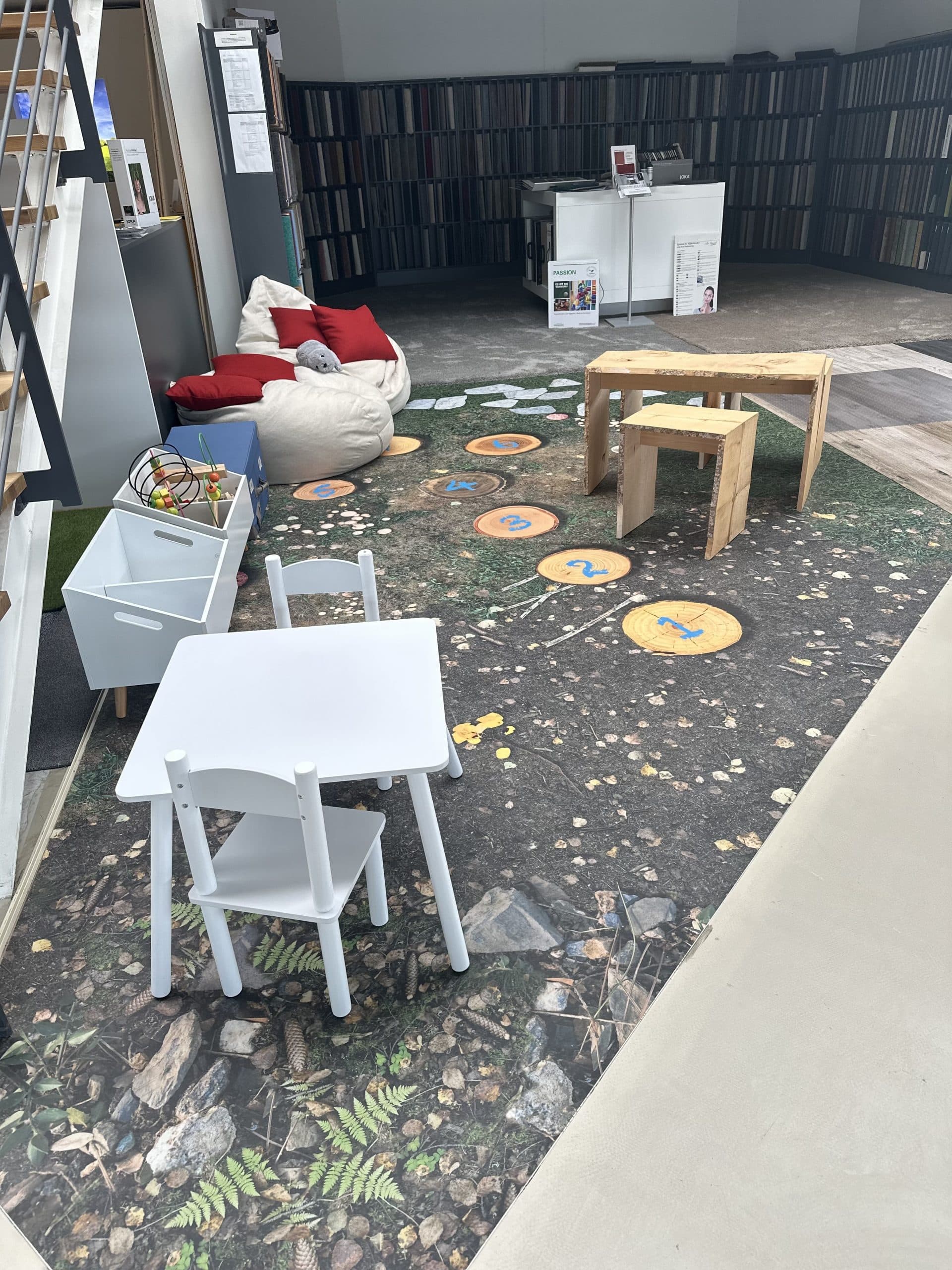 Der Raum zeigt eine kindgerechte Ecke mit einem weißen Tisch und einem Stuhl, umgeben von einem bunten Bodenbelag, der Naturmotive darstellt. Es gibt eine gemütliche Sitzgelegenheit aus einem großen, beigen Kissen mit roten Kissen, sowie ein Holzspielzeug und eine Box mit weiteren Spielmaterialien. Im Hintergrund sind Regale mit verschiedenen Materialien zu sehen.