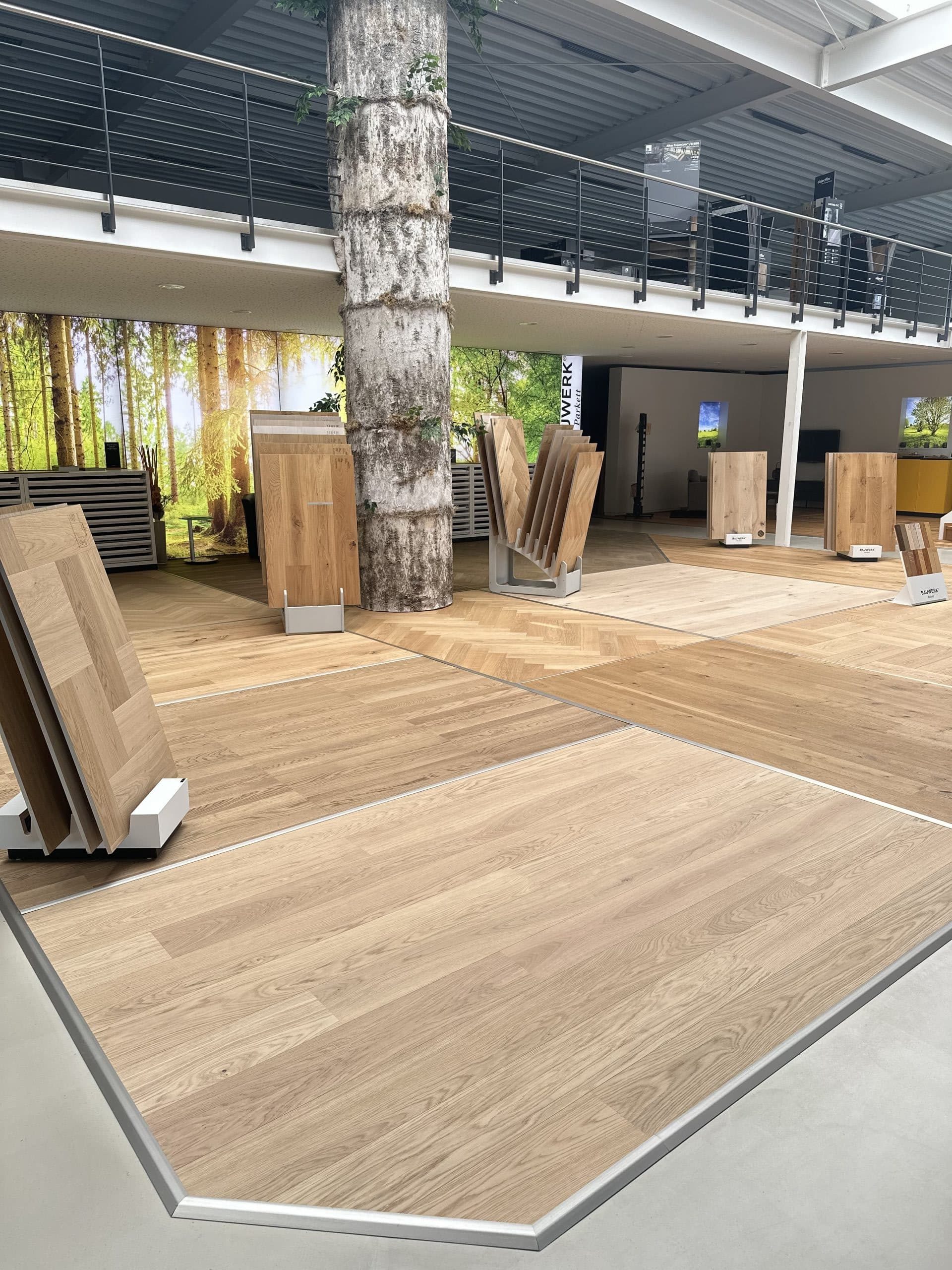 Ein Ausstellungsraum mit Holzfußbodenmustern und einem großen Baum in der Mitte. Im Hintergrund sind Bilder von Wäldern zu sehen, während verschiedene Holzpaneele in verschiedenen Stilen und Farben präsentiert werden.