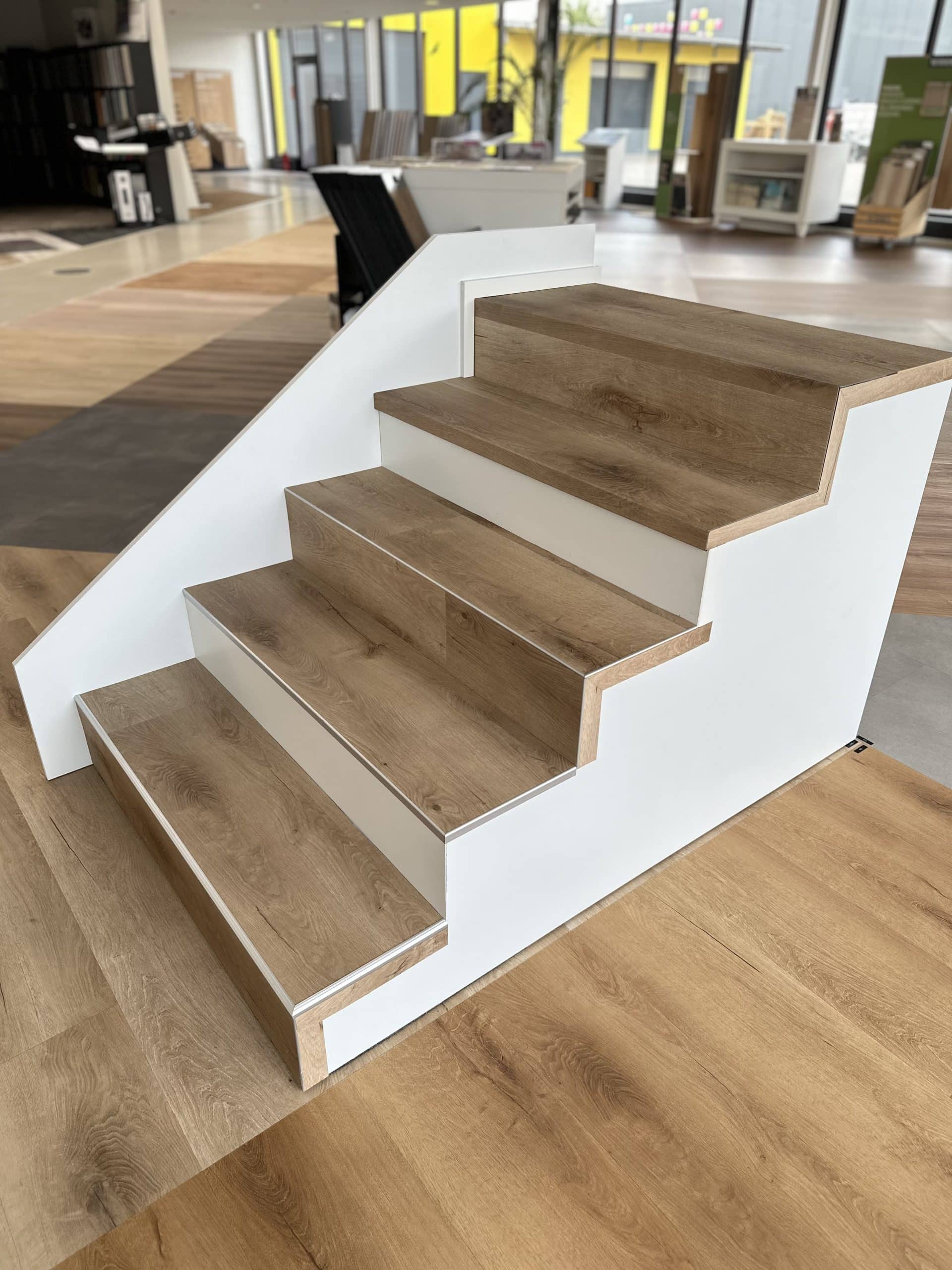 Eine moderne Treppe mit Holzstufen und einem weißen Geländer, umgeben von einem hellen Innenraum mit verschiedenen Bodenbelägen und großen Fenstern.