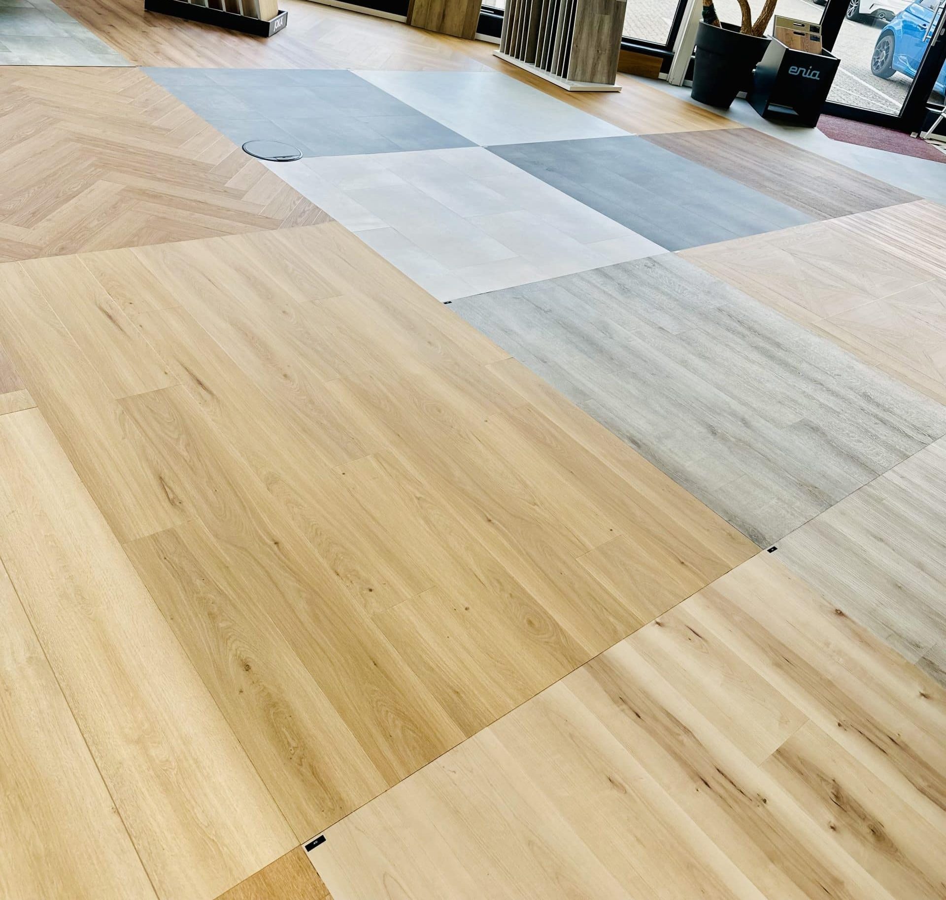 Der Boden besteht aus verschiedenen Laminat- und Holzplatten in unterschiedlichen Farbtönen und Mustern, die in einem modernen Raum verlegt sind. Einige Bereiche zeigen helle Holzoptik, während andere in Grau- und Beigetönen gehalten sind.