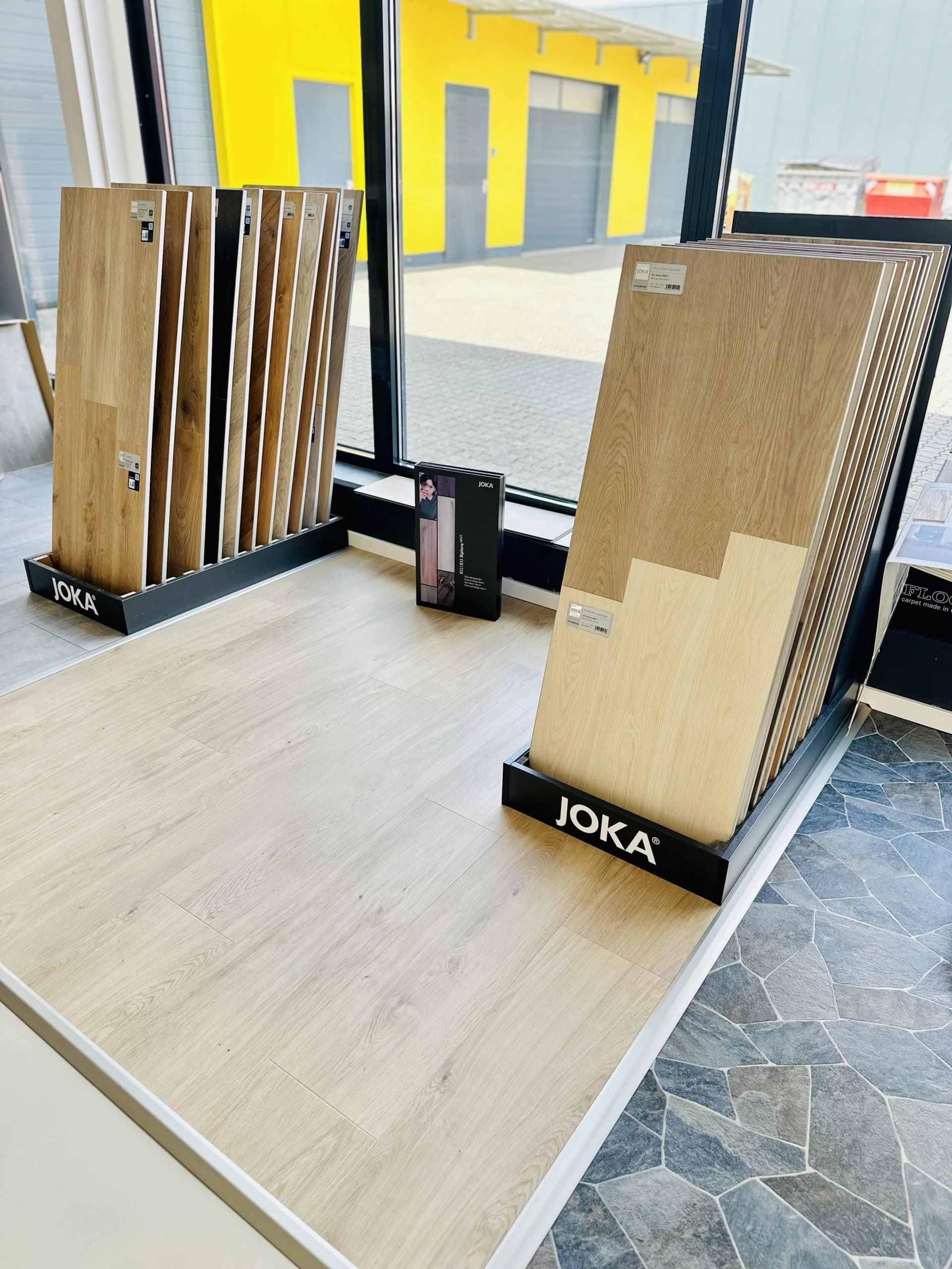 In einem modernen Ausstellungsraum stehen verschiedene Bodenbeläge der Marke JOKA. Auf der linken Seite sind Holzpaneele in verschiedenen Farben aufgestellt, während auf dem Boden ein heller, holzähnlicher Belag verlegt ist. Daneben liegt eine Produktbroschüre von JOKA. Im Hintergrund sind große Fenster, die viel Tageslicht hereinlassen, und eine gelbe Wand ist sichtbar.