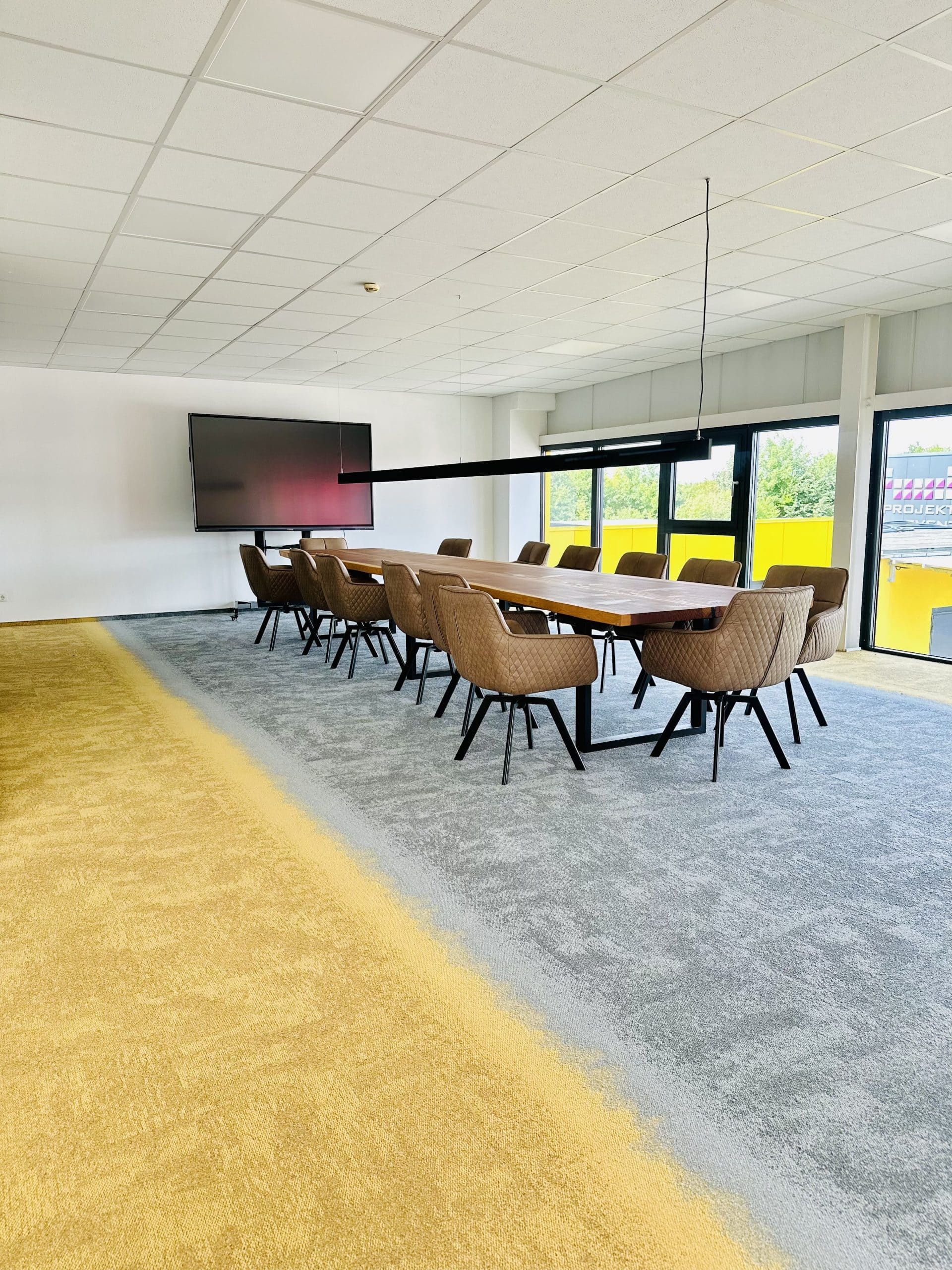 Ein moderner Konferenzraum mit einem großen Tisch und mehreren Stühlen, ausgestattet mit einem großen Bildschirm an der Wand. Der Boden ist in zwei Farben gestaltet, mit einem grauen und einem gelben Teppichbereich. Große Fenster sorgen für viel Tageslicht.