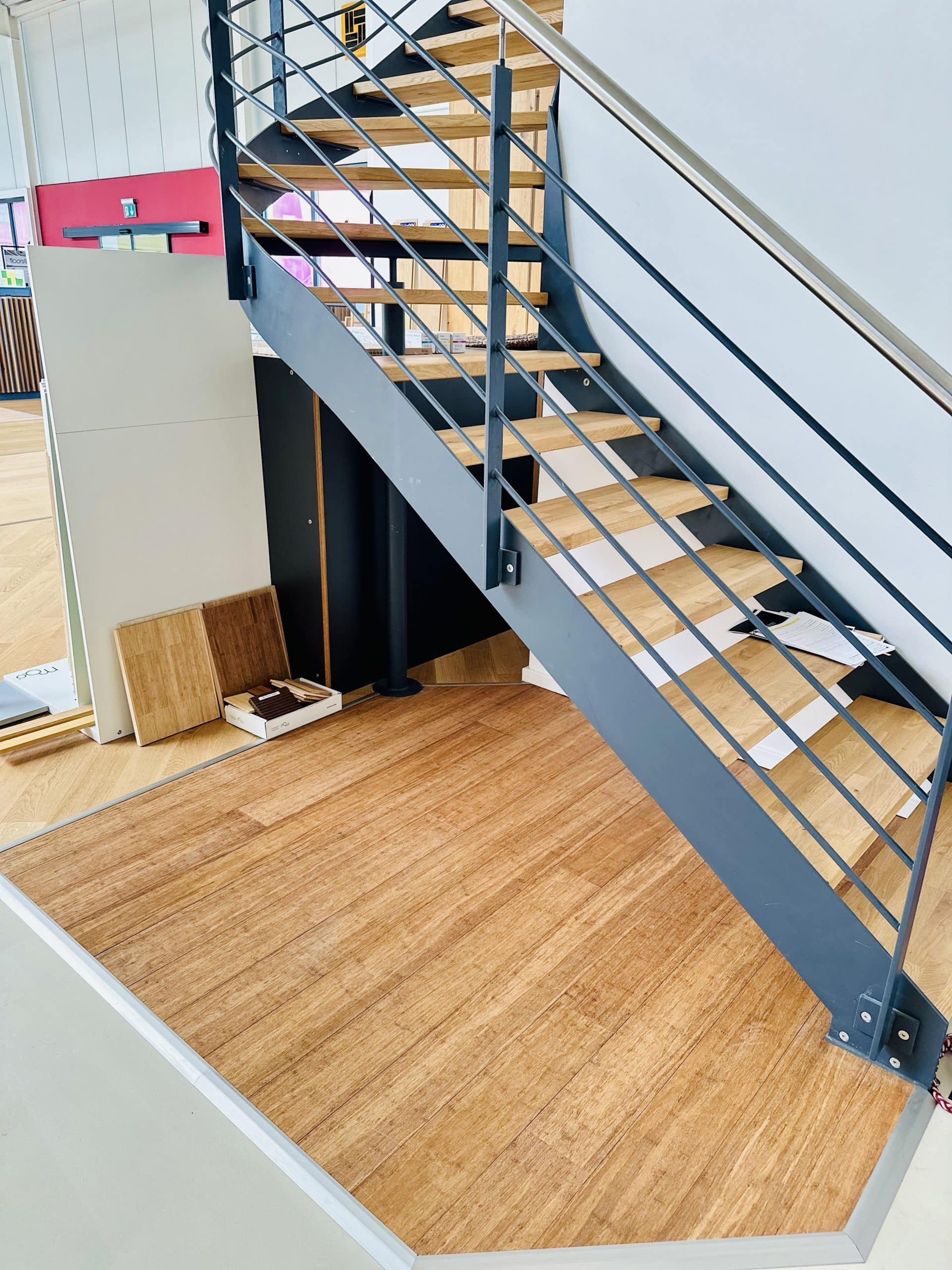 Eine moderne Treppe mit Holzstufen und einem Stahlgeländer, die in einem hellen Raum steht. Unter der Treppe befinden sich verschiedene Bodenbeläge, darunter Holzplatten und einige Materialien zur weiteren Gestaltung.
