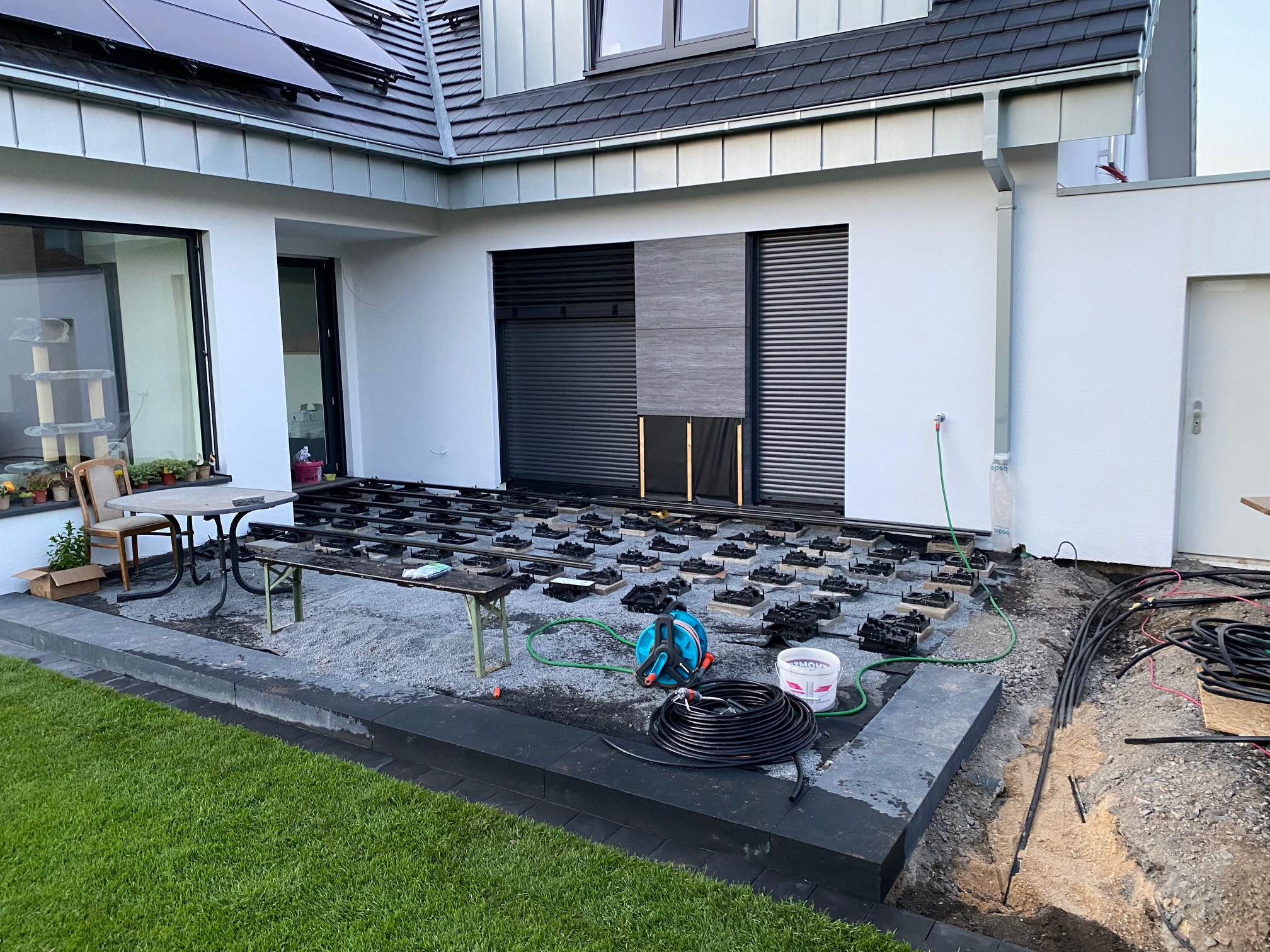 Blick auf eine Baustelle im Außenbereich eines modernen Hauses, wo der Boden mit verschiedenen Materialien vorbereitet wird. Im Vordergrund steht ein Tisch mit einem Stuhl, während im Hintergrund eine Wand mit Rollladen und Solarpanelen sichtbar ist. Auf dem Boden liegen mehrere schwarze Platten und Rohre sind verlegt.