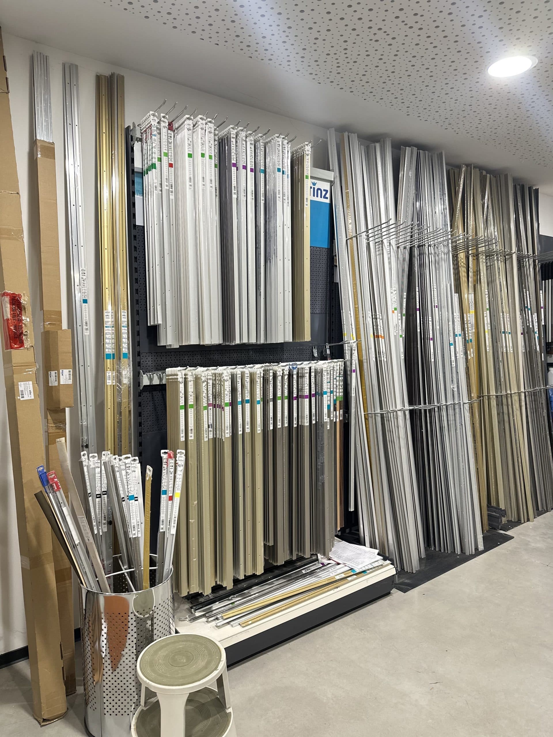 Ein Regal mit verschiedenen Aluminiumprofilen und -stangen in unterschiedlichen Längen und Farben, die ordentlich aufgereiht sind. Im Vordergrund steht ein runder Metallbehälter mit weiteren Profilen und daneben ein kleiner Hocker. Der Boden ist aus grauem Beton.