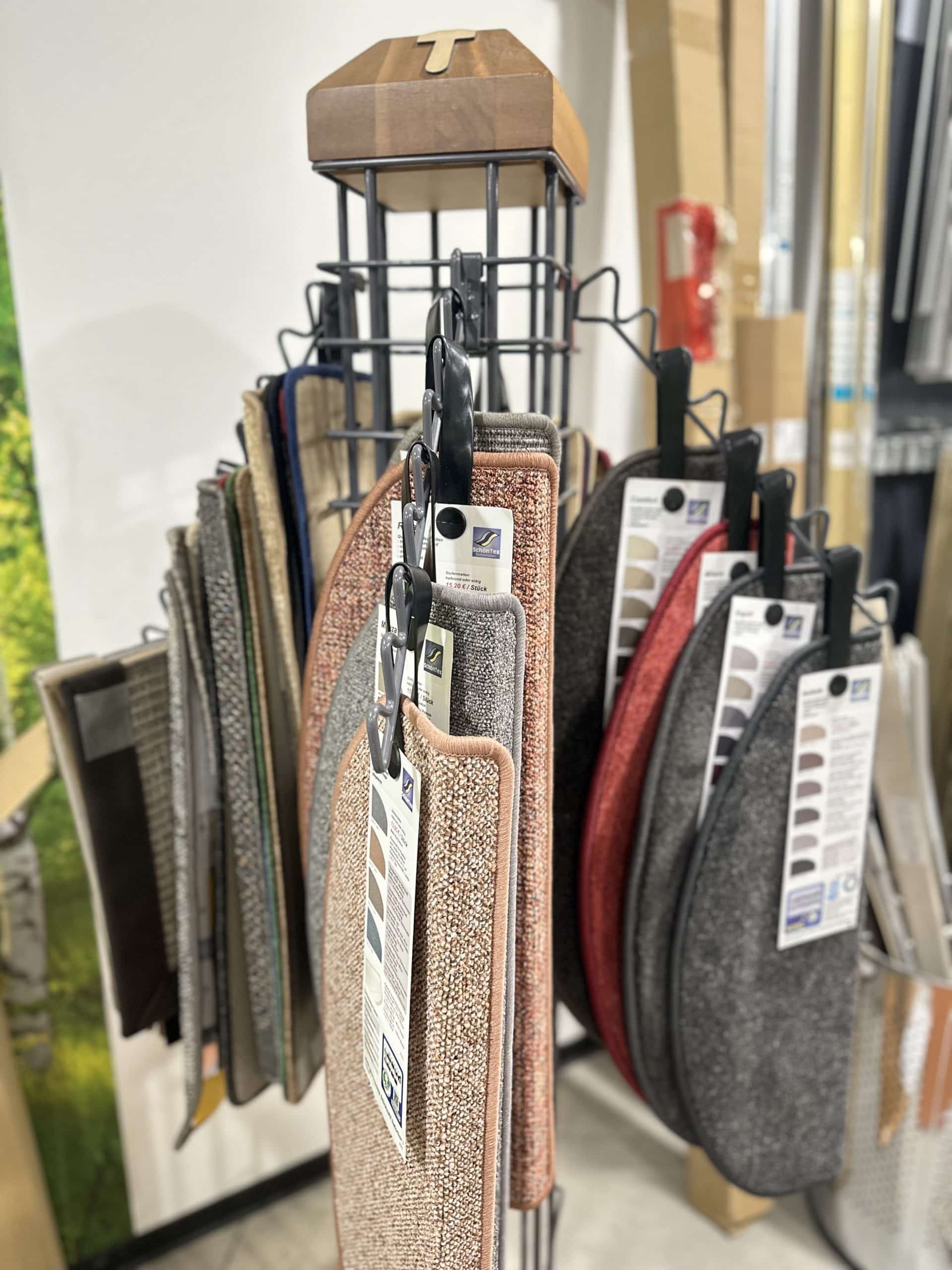 Ein Ständer mit verschiedenen Teppichmustern hängt in einem Geschäft, die Muster sind in verschiedenen Farben und Texturen angeordnet. Oben befindet sich ein Holzteil mit einem Buchstaben 