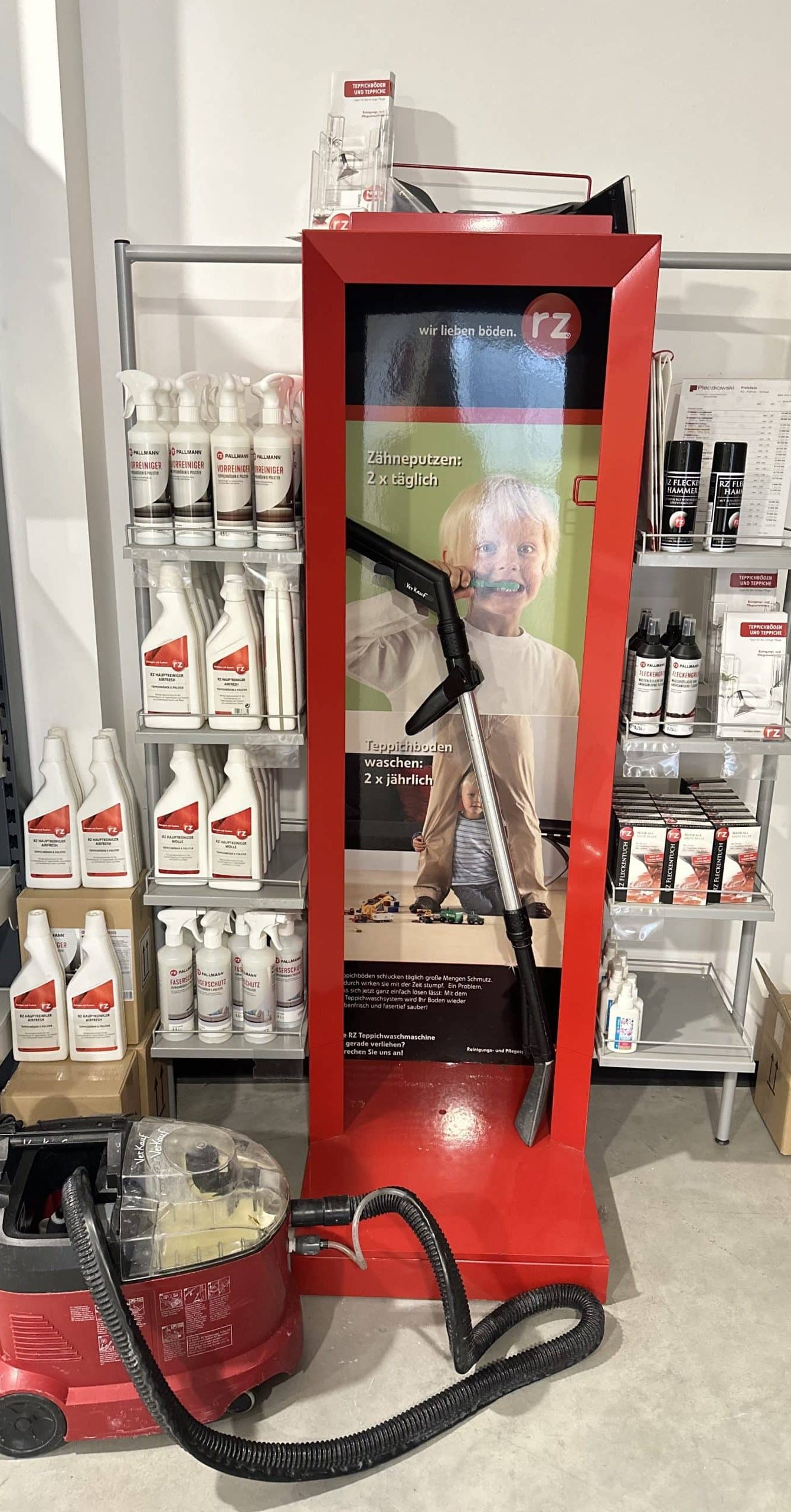 Ein Verkaufsdisplay mit einem Kind, das Zähne putzt, und einem Teppichreiniger im Vordergrund. Im Hintergrund sind verschiedene Reinigungsmittel in weißen Flaschen auf einem Regal angeordnet. Das Display enthält Informationen über die Reinigung von Teppichböden und zeigt, dass der Teppich mindestens zweimal jährlich gewaschen werden sollte.