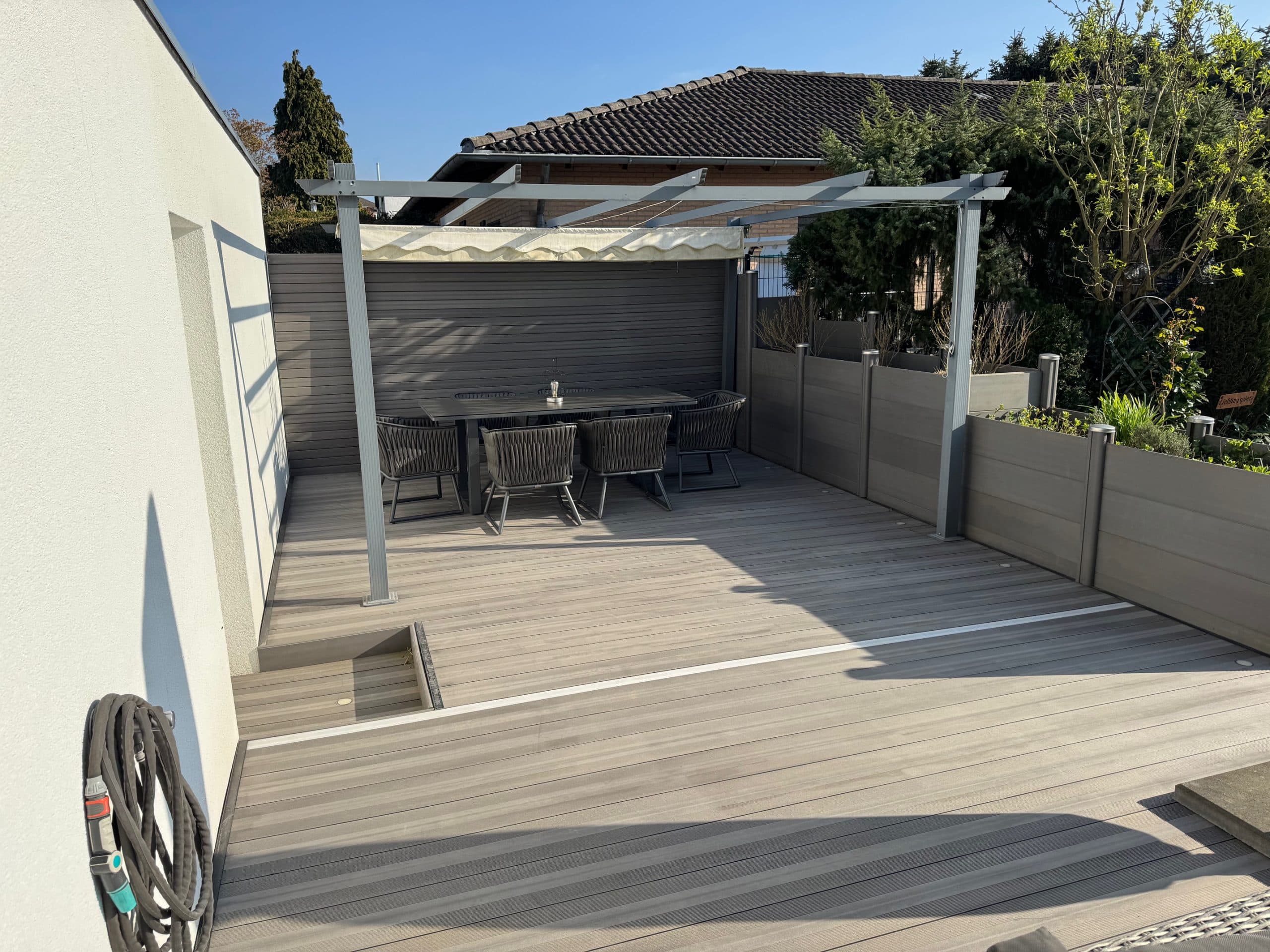 Eine moderne Terrasse mit einem Holzboden, einem großen Tisch und Stühlen, umgeben von niedrigen Wänden und Pflanzen. Über dem Tisch befindet sich eine Markise, und der Himmel ist klar und blau.