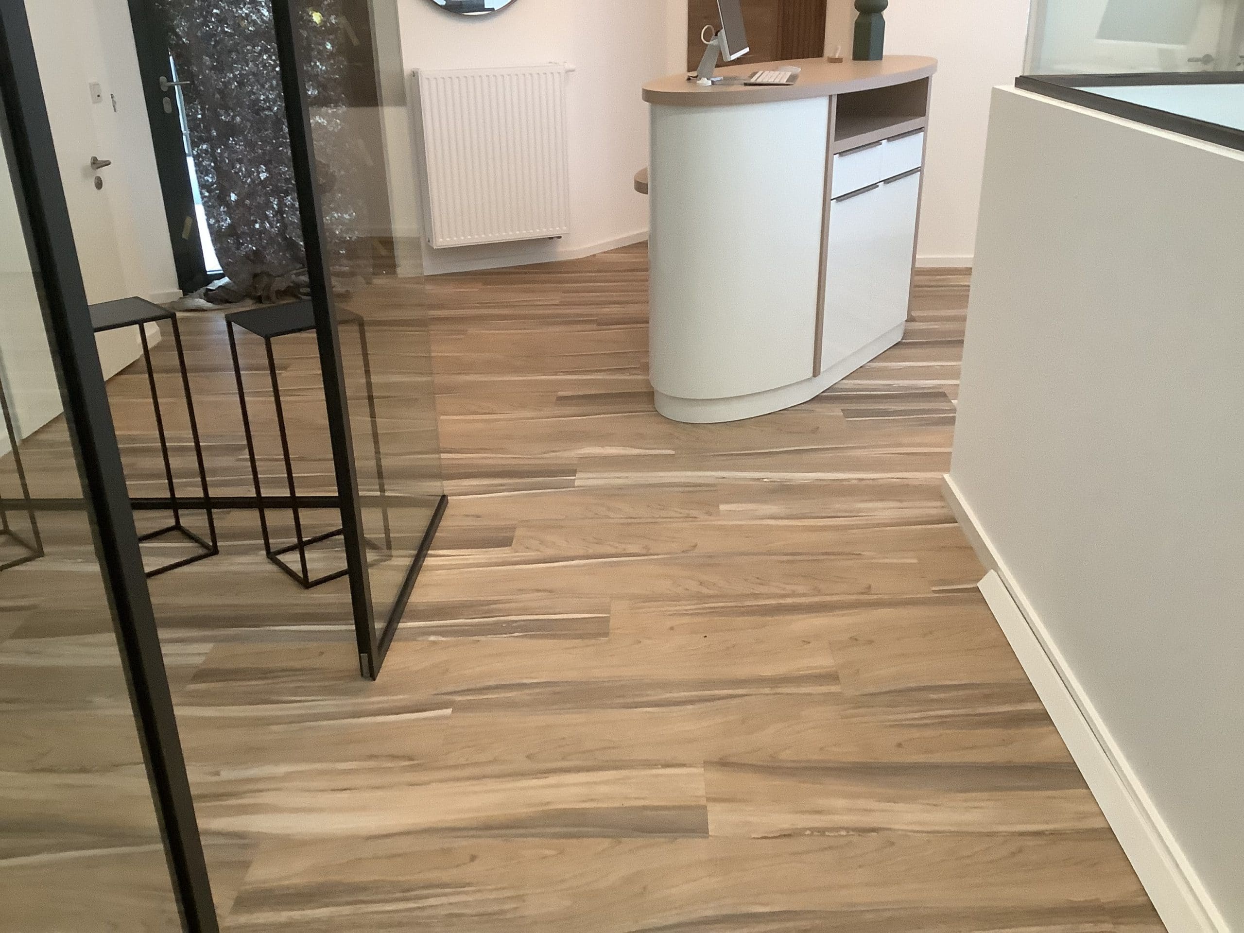 Ein moderner Empfangsbereich mit einem hellen Holzboden, einem geschwungenen Empfangstresen und zwei hohen, schwarzen Hockern. Im Hintergrund sind große Fenster und eine Heizung sichtbar.