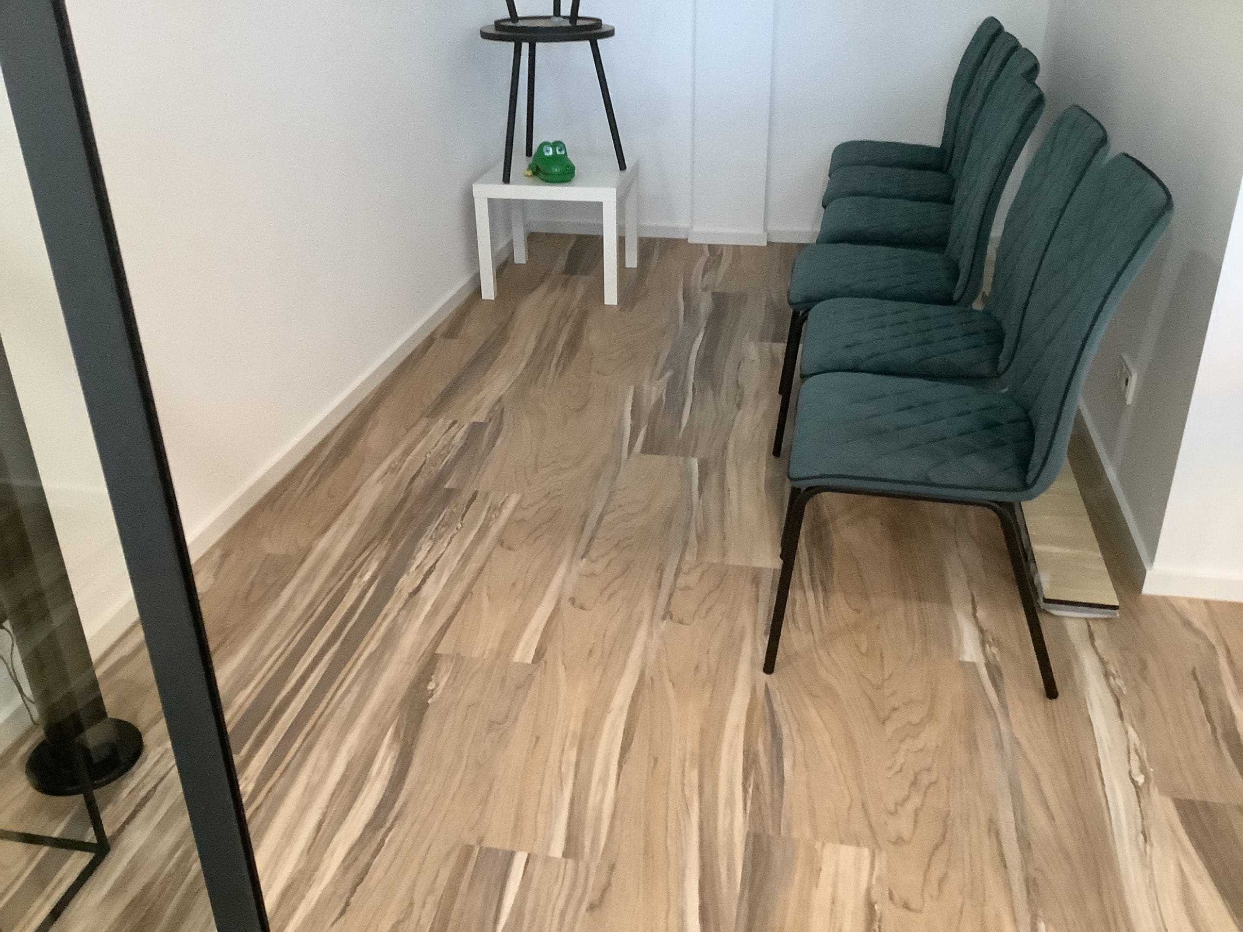 Ein heller Raum mit einem modernen, holzähnlichen Bodenbelag. An der Wand stehen fünf grüne Stühle mit gepolsterten Rückenlehnen. Neben den Stühlen befindet sich ein kleiner, weißer Tisch mit einem grünen Dekorationsobjekt darauf.