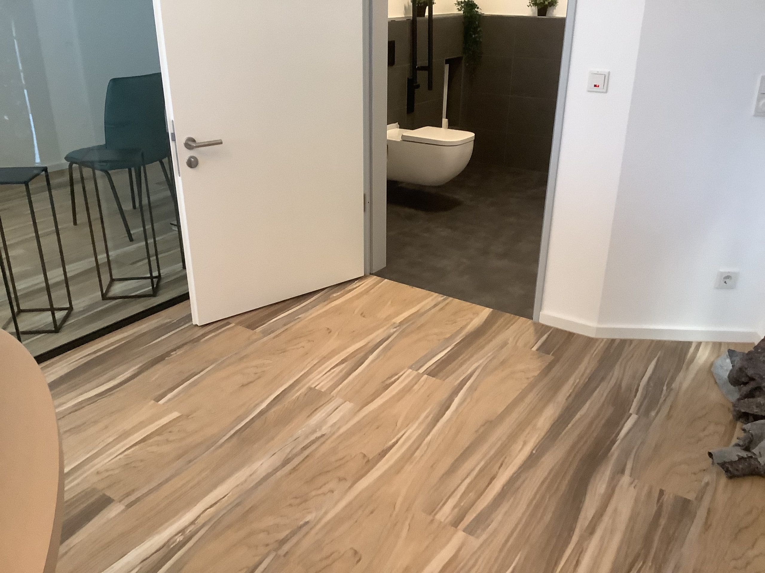 Der Boden ist aus hellem Holz mit einer auffälligen Maserung, und eine offene Tür führt in einen modernen, gefliesten Raum mit einer freistehenden Toilette. Im Hintergrund sind stilvolle, hohe Stühle und ein Tisch zu sehen.