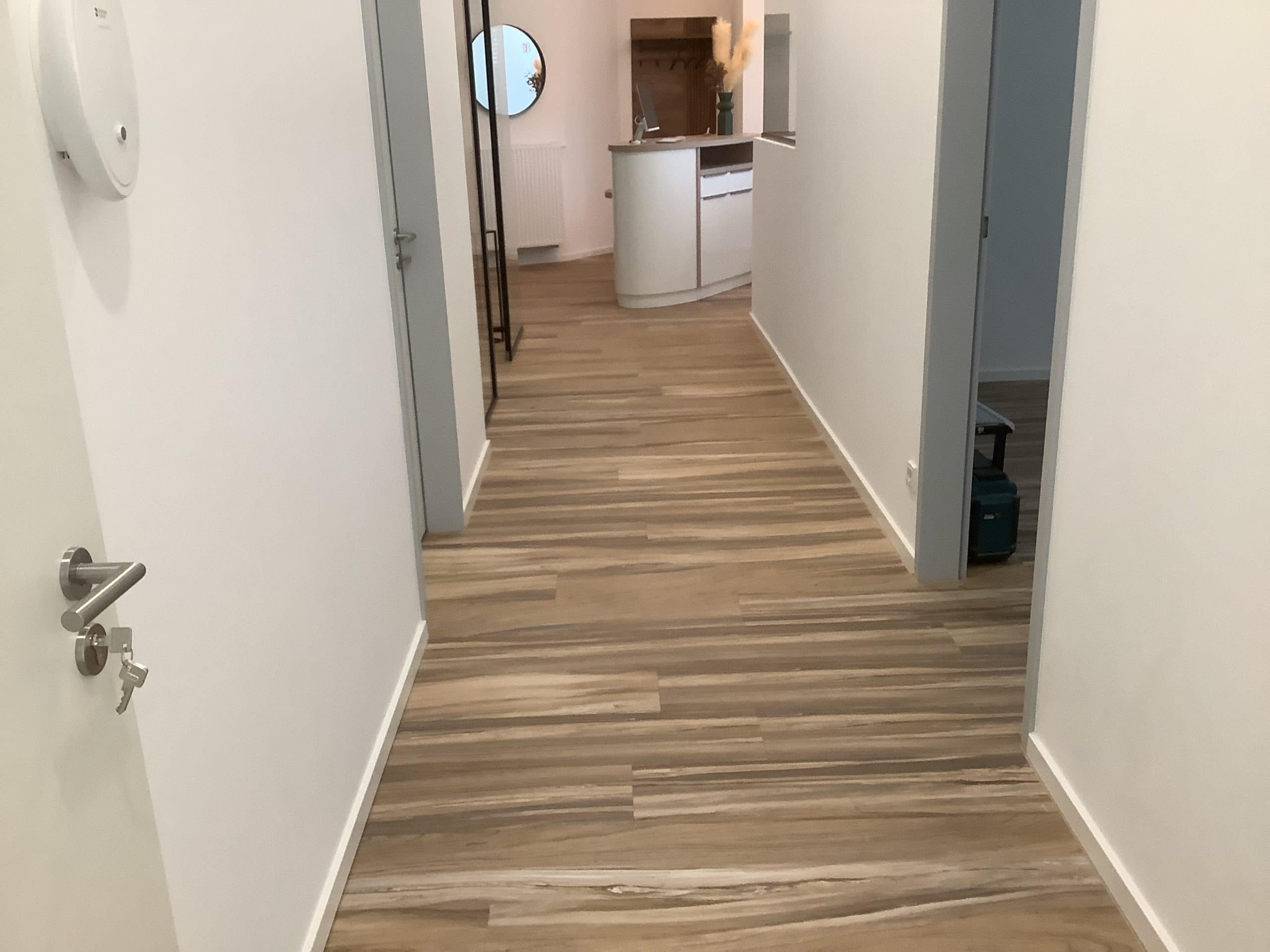 Ein heller Flur mit Holzfußboden, der zu einem offenen Wohnbereich führt. An der Wand sind Türen zu sehen, und in der Ferne ist eine moderne Küche mit einer runden Theke sichtbar. Ein runder Spiegel hängt an der Wand, und es gibt eine kleine Pflanze auf der Theke.