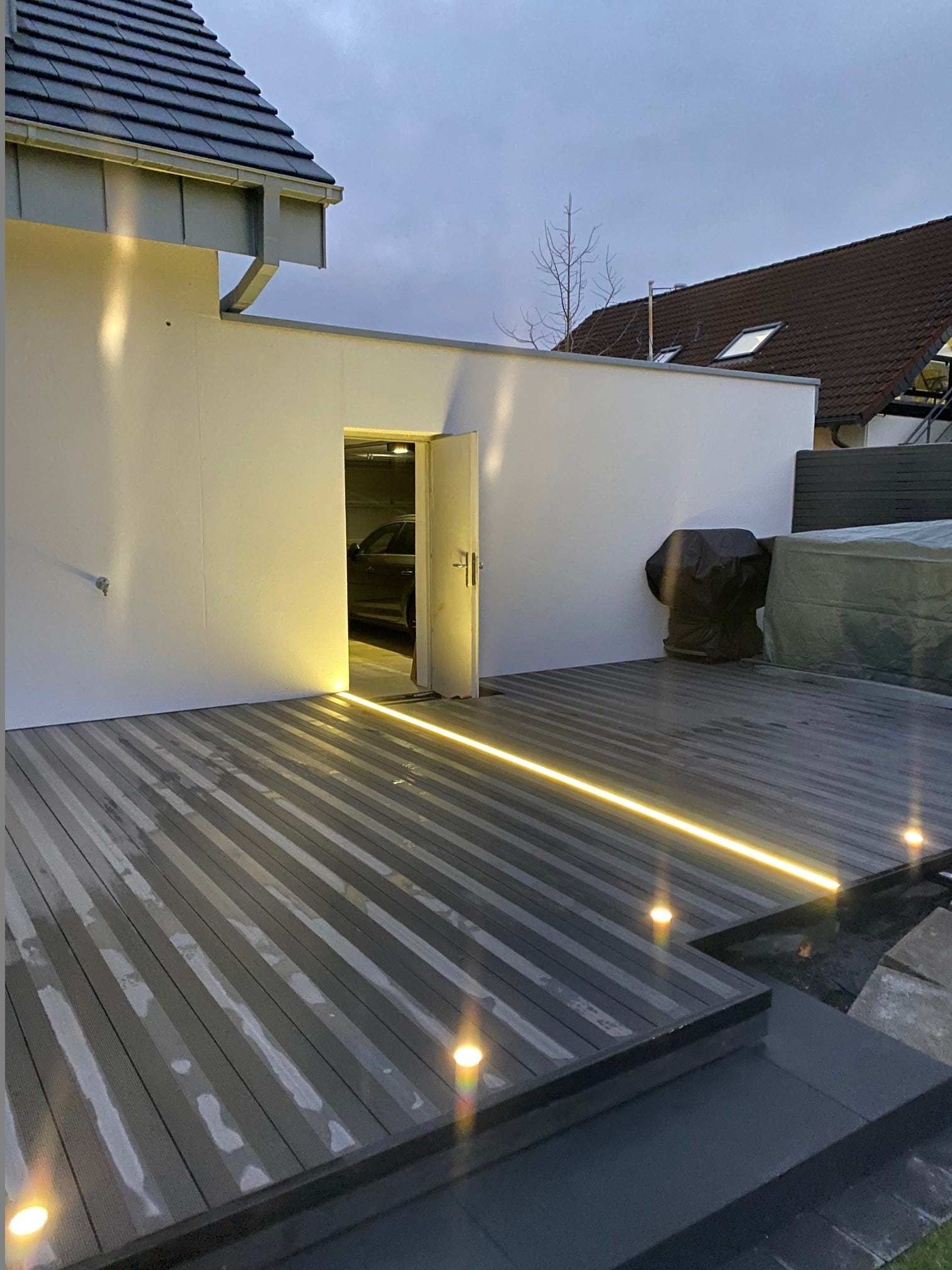 Eine moderne Terrasse mit Holzplanken, die sanft beleuchtet ist. Im Hintergrund ist eine offene Tür zu sehen, die in einen Garagenbereich führt. Der Himmel ist bewölkt und es ist Dämmerung.