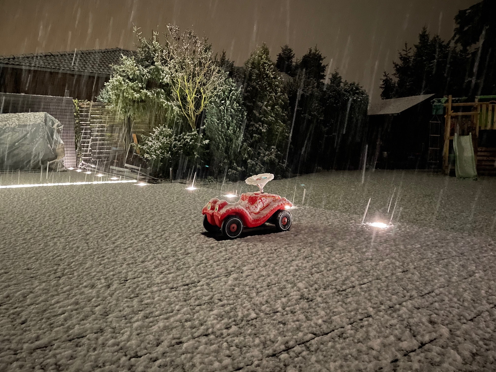 Ein rotes Spielzeugauto steht alleine auf einem schneebedeckten Gartenboden, während es stark schneit. Im Hintergrund sind Bäume und eine Gartenhütte zu sehen, die von sanftem Licht beleuchtet werden.
