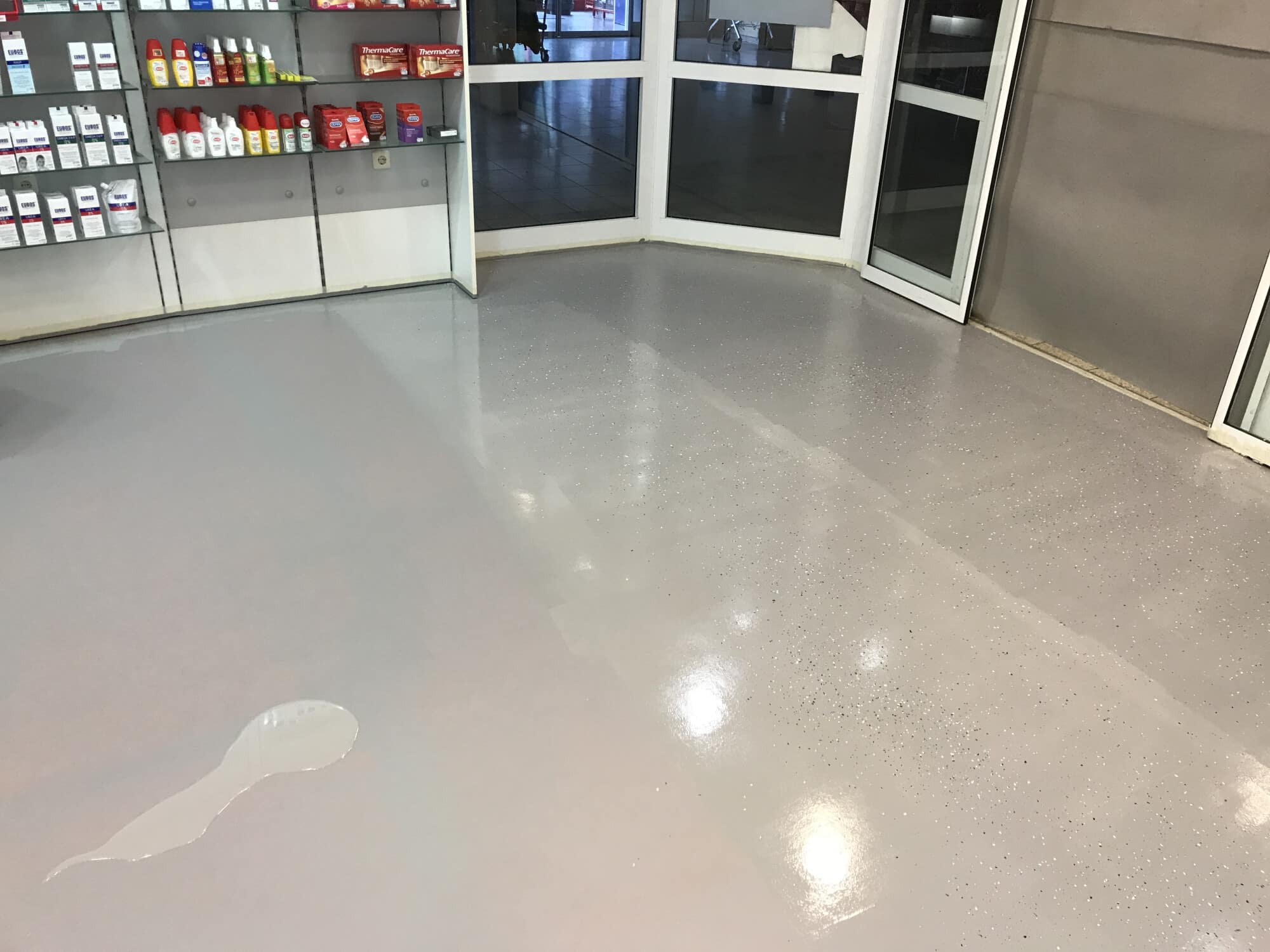 Ein glatter, grauer Boden in einem Verkaufsraum, in dem verschiedene Pflegeprodukte in Regalen ausgestellt sind. An einer Stelle ist eine kleine Pfütze sichtbar. Die Eingangstür ist offen und lässt Tageslicht herein.