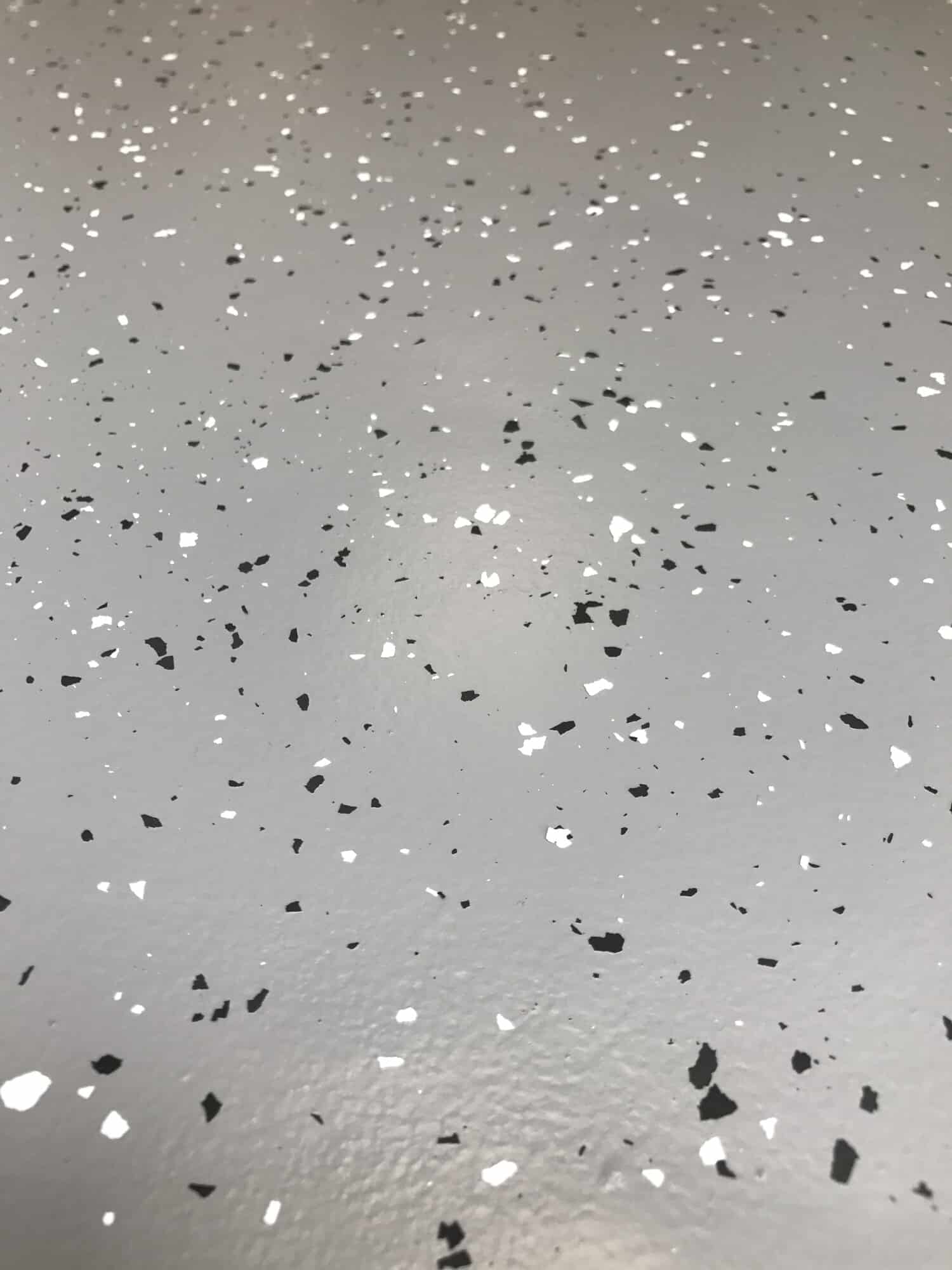 Eine graue Bodenfläche mit schwarzen und weißen Farbspänen, die gleichmäßig verteilt sind und eine strukturierte Oberfläche bilden.