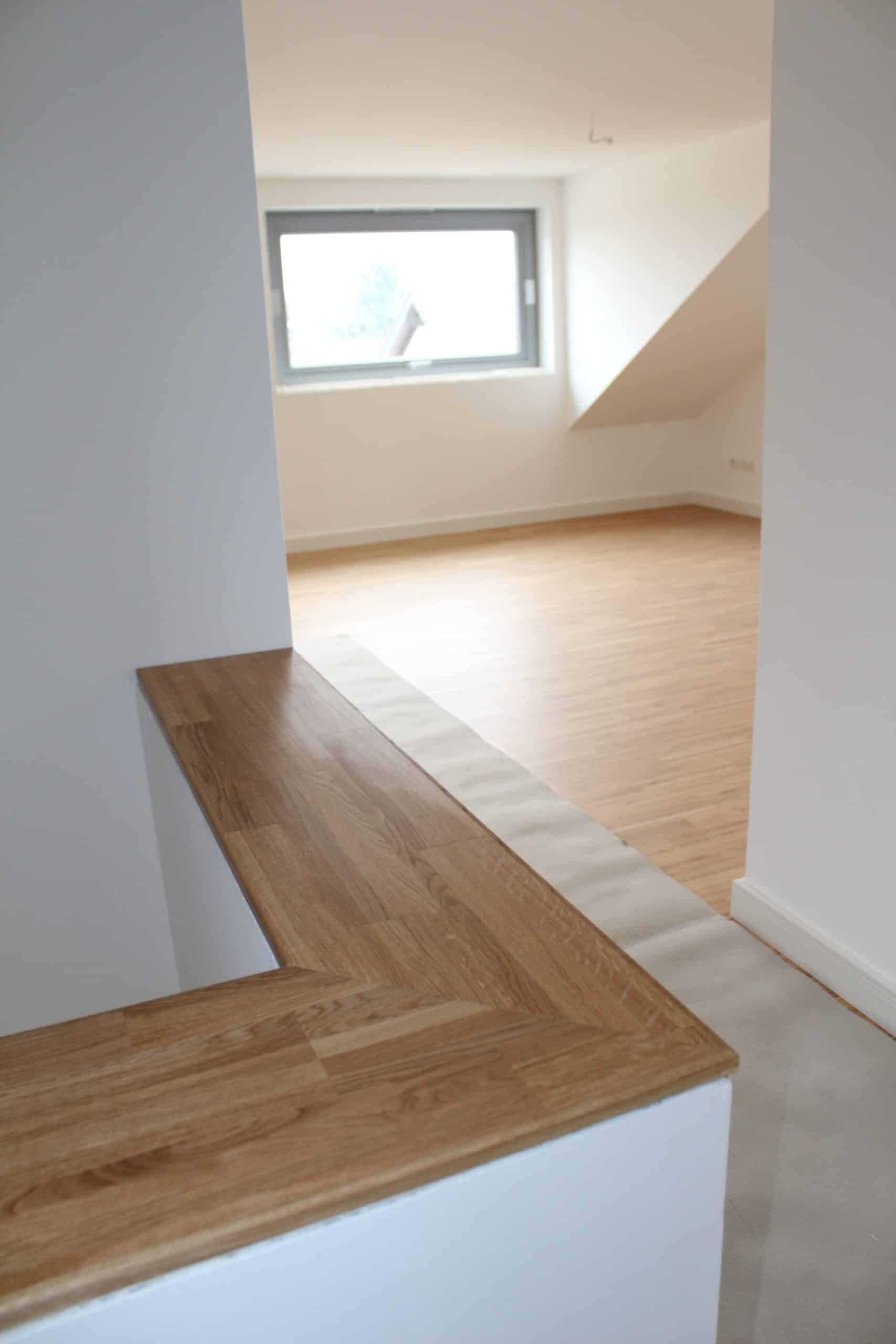 Ein heller Raum mit einem großen Fenster, das Tageslicht hereinlässt. Der Boden ist mit hellem Holz ausgelegt, und im Vordergrund ist eine Holzablage zu sehen, die an einer Wand angrenzt. Die Wände sind schlicht und weiß, was dem Raum eine moderne und offene Atmosphäre verleiht.