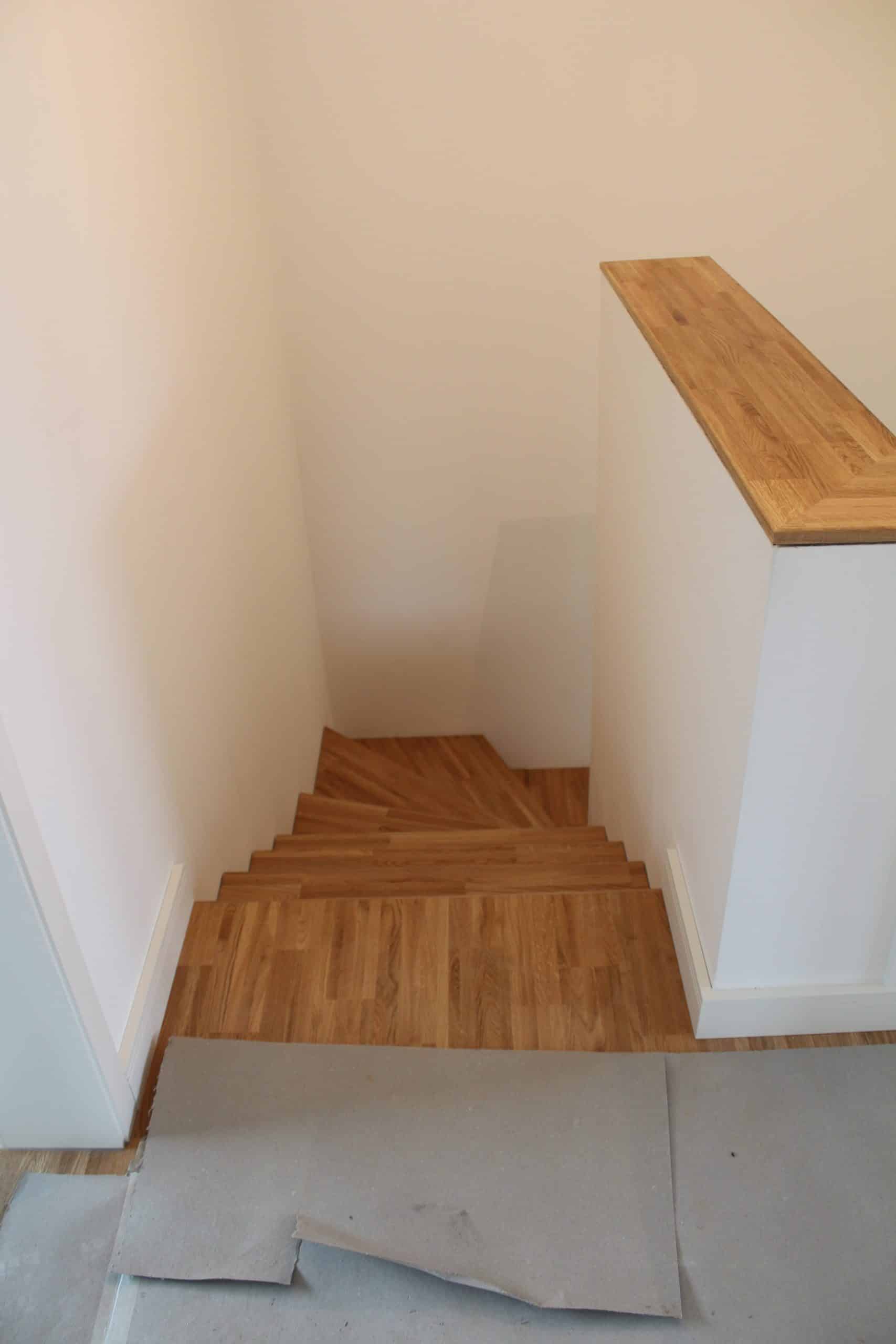 Eine Treppe aus Holz, die in einen hellen, weißen Raum führt. Der Boden ist ebenfalls mit Holz ausgelegt, und auf einer Stufe liegt ein graues Material, das möglicherweise zum Schutz während der Renovierung dient.