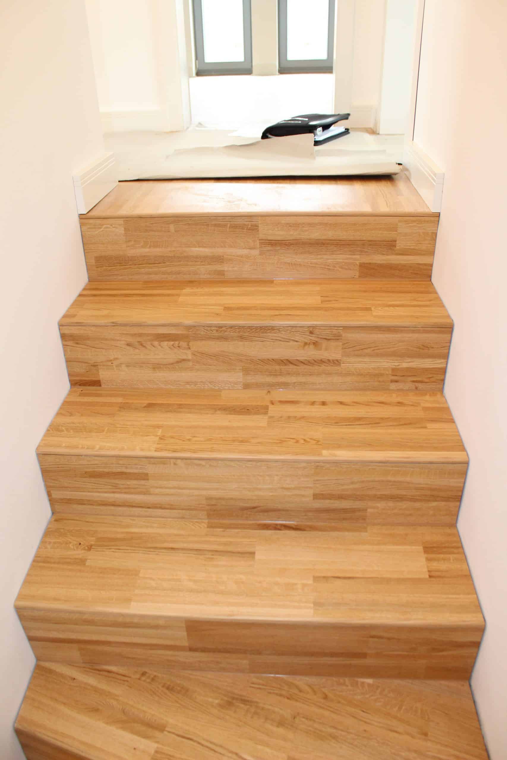 Eine Treppe mit hellen Holzstufen, die zu einem oberen Bereich führt. Im Hintergrund sind Fenster zu sehen, die viel Tageslicht hereinlassen.