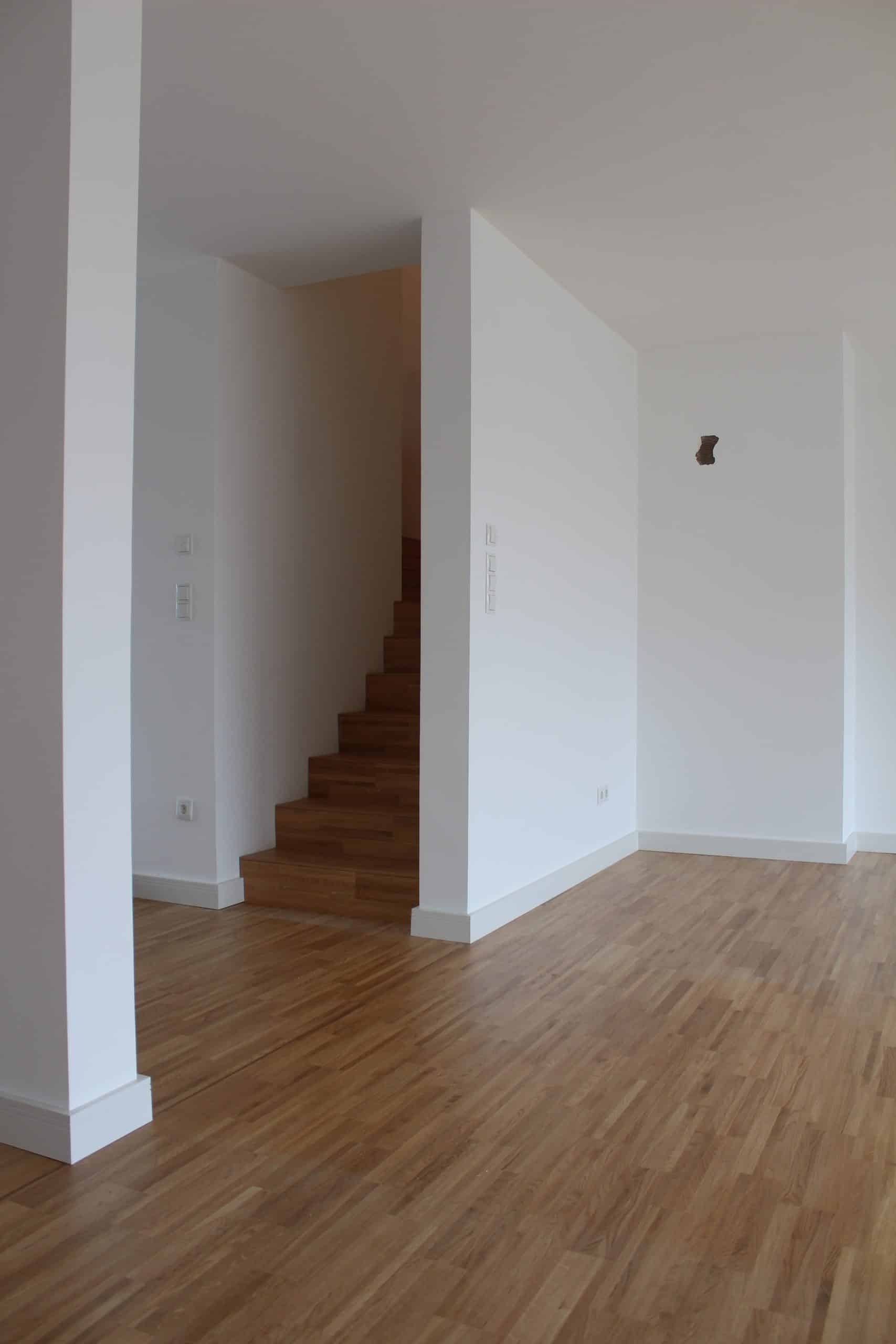 Ein heller, minimalistischer Raum mit Holzfußboden, weißen Wänden und einer Treppe im Hintergrund.