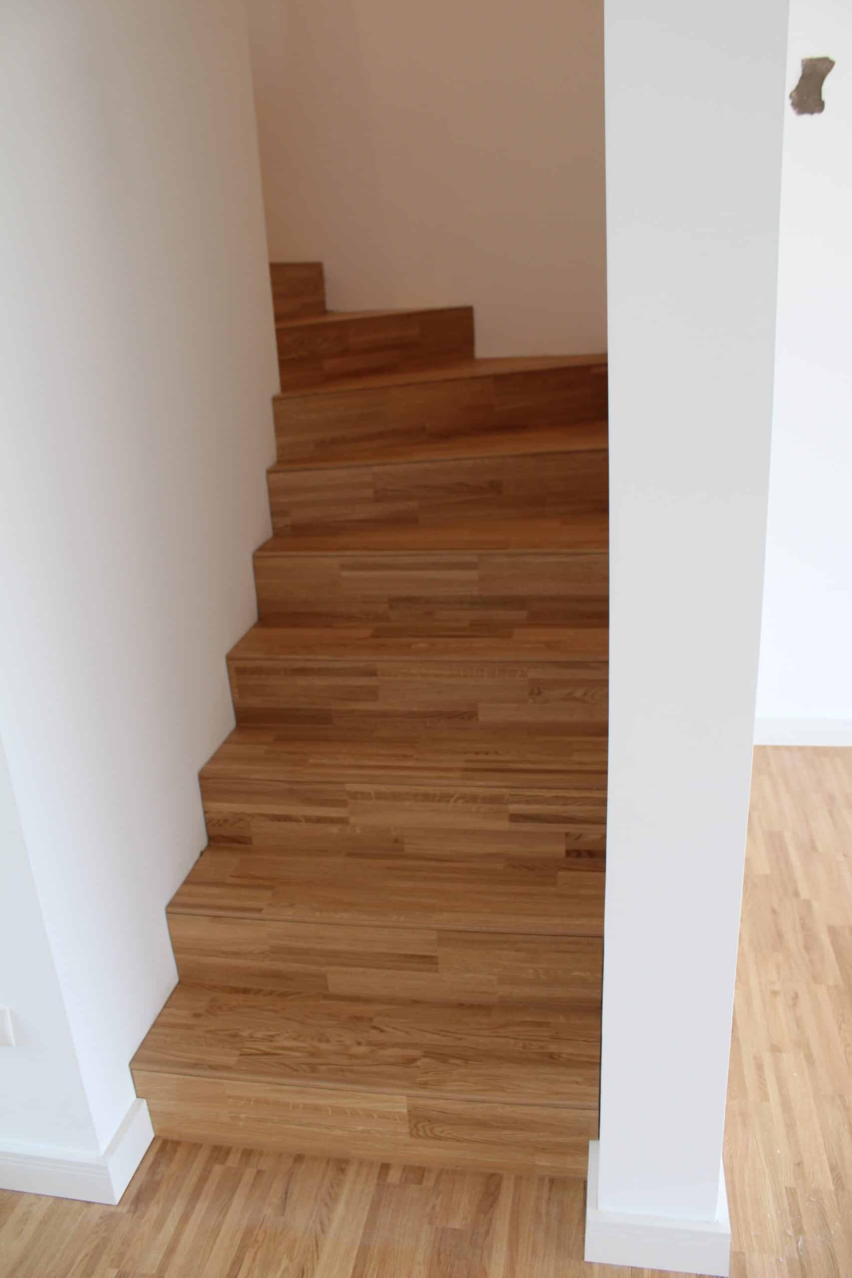 Eine Treppe aus Holz mit hellen Stufen, die in einem modernen Innenraum verläuft. Die Wände sind weiß gestrichen, und der Boden besteht aus hellen Holzpaneelen.