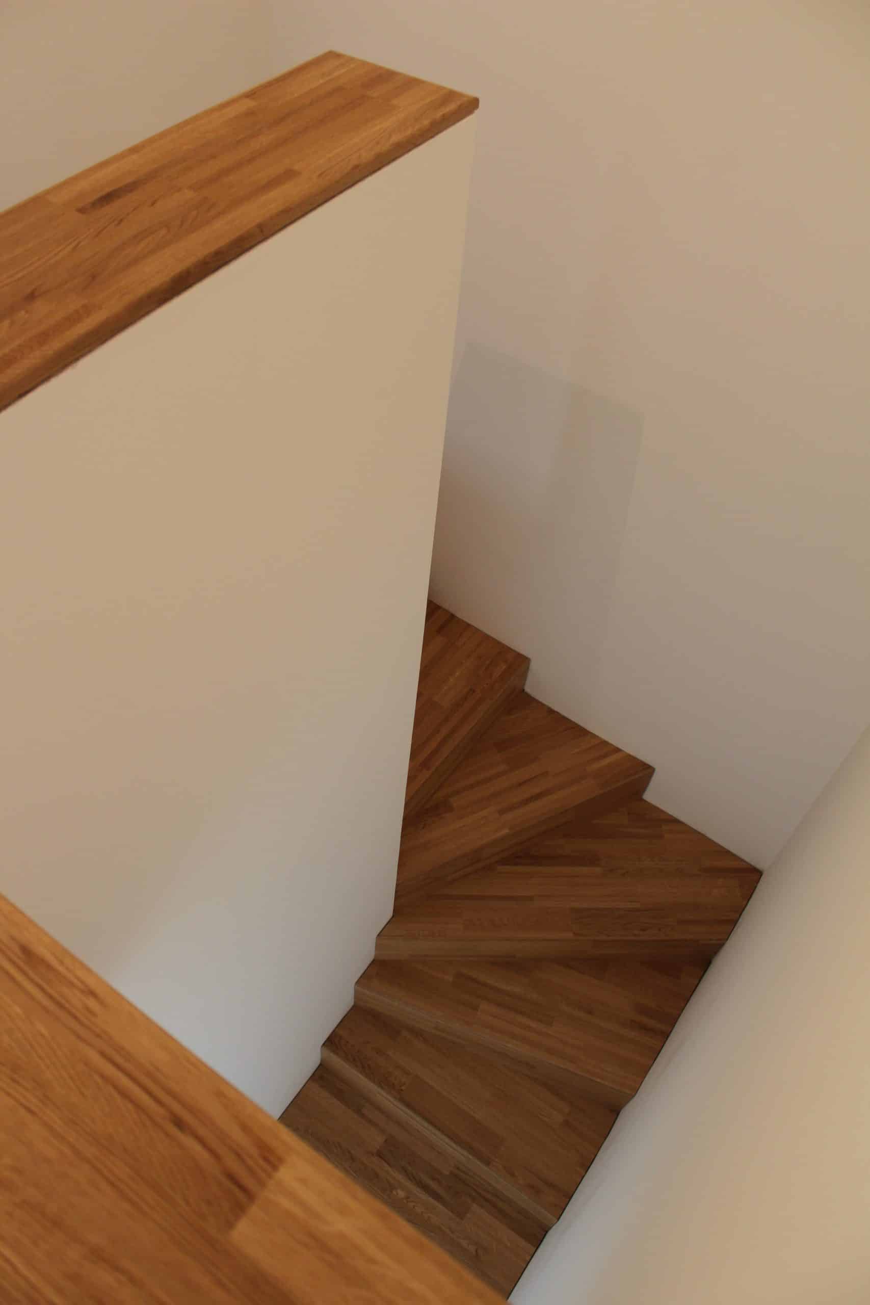 Eine spiralförmige Holztreppe, die in einem hellen Raum mit weißen Wänden verläuft. Der Blick von oben zeigt die geschwungenen Stufen aus Holz und ein Geländer aus Holz.